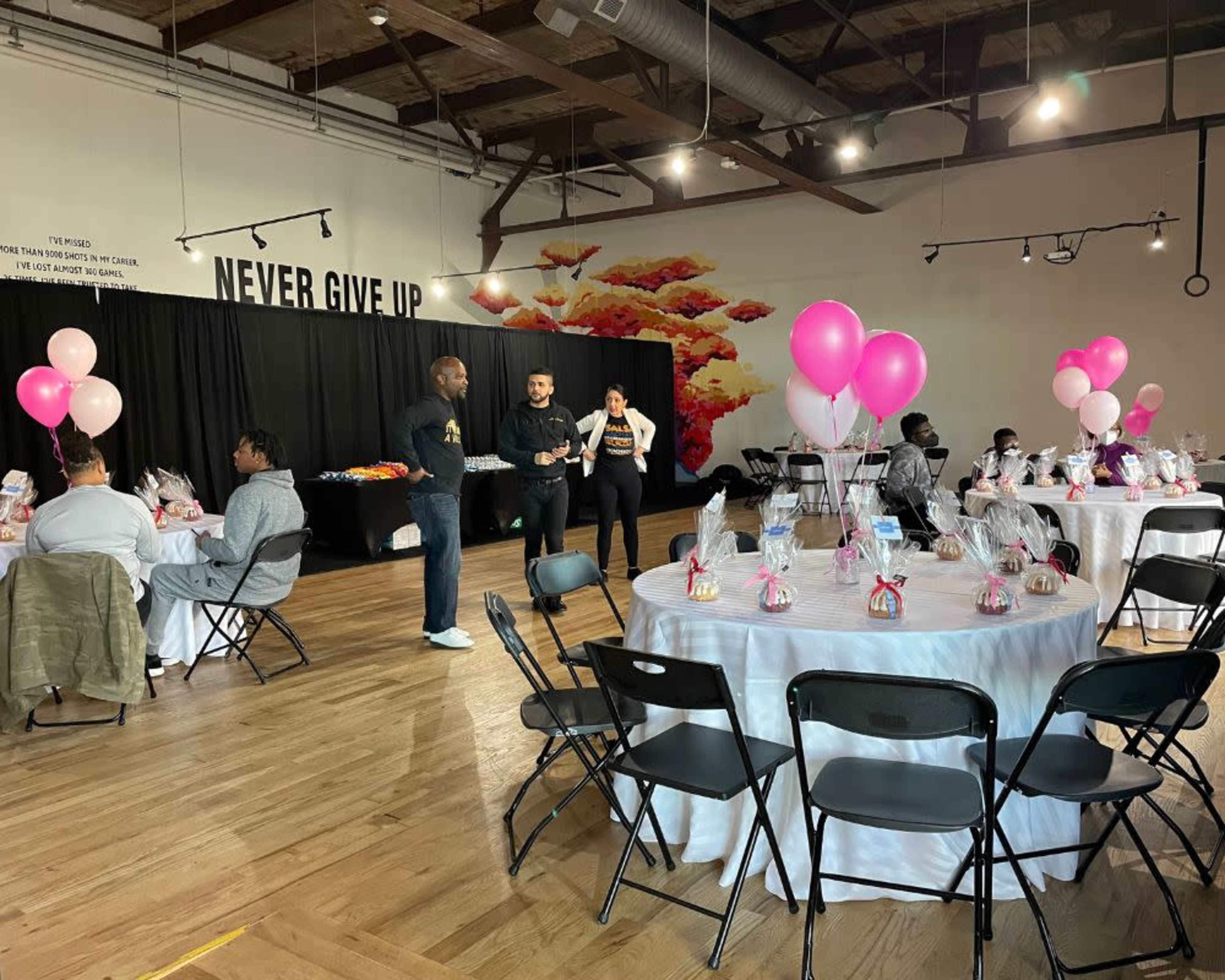 A spacious event hall is set up for a gathering, featuring round tables with party favors and balloons, while guests interact and staff members assist.