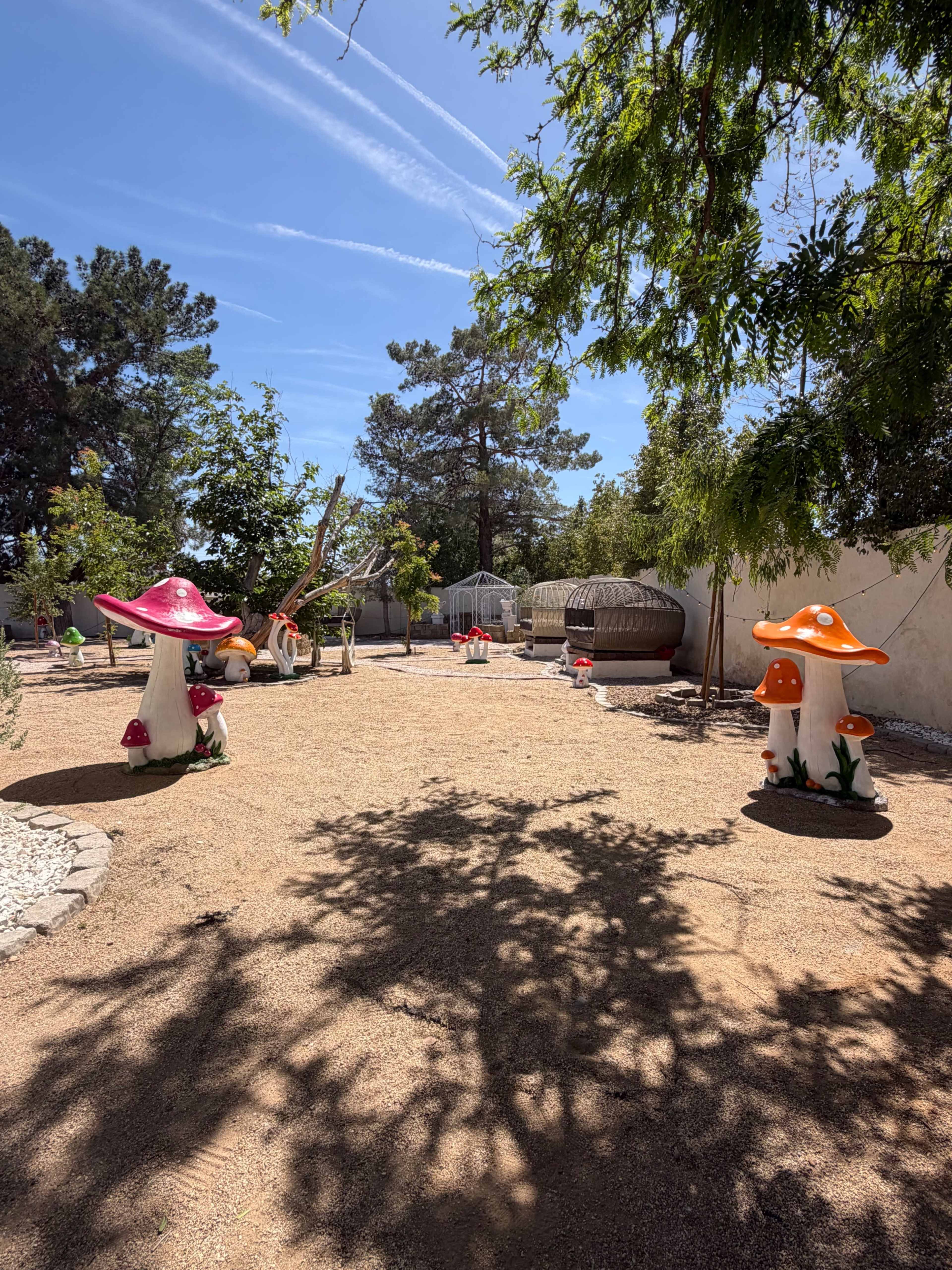 The image features a garden scene with decorative oversized mushroom sculptures amidst gravel pathways and various trees under a clear blue sky.