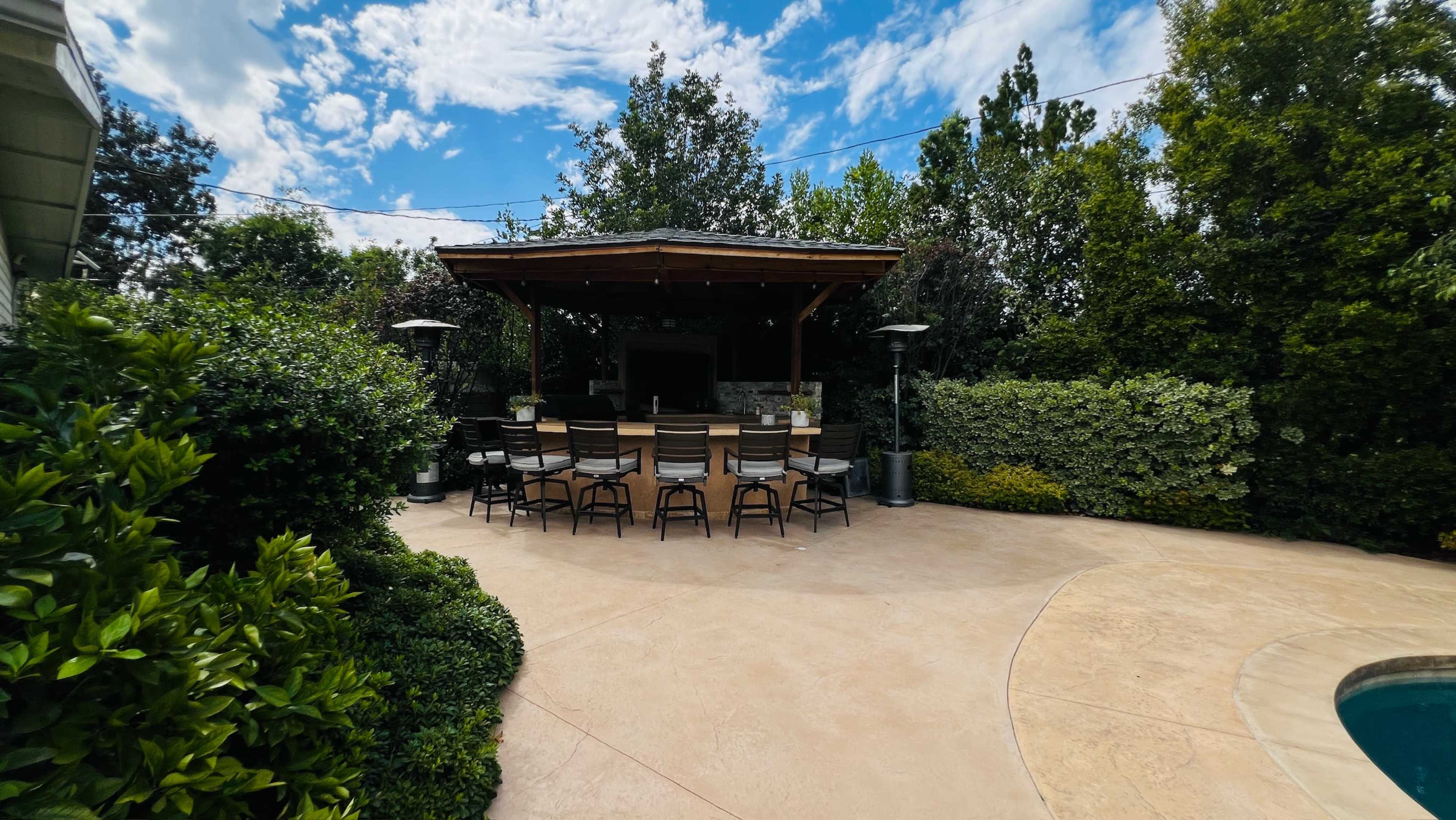 A patio area features a large table with multiple chairs under a wooden gazebo surrounded by greenery and a swimming pool.