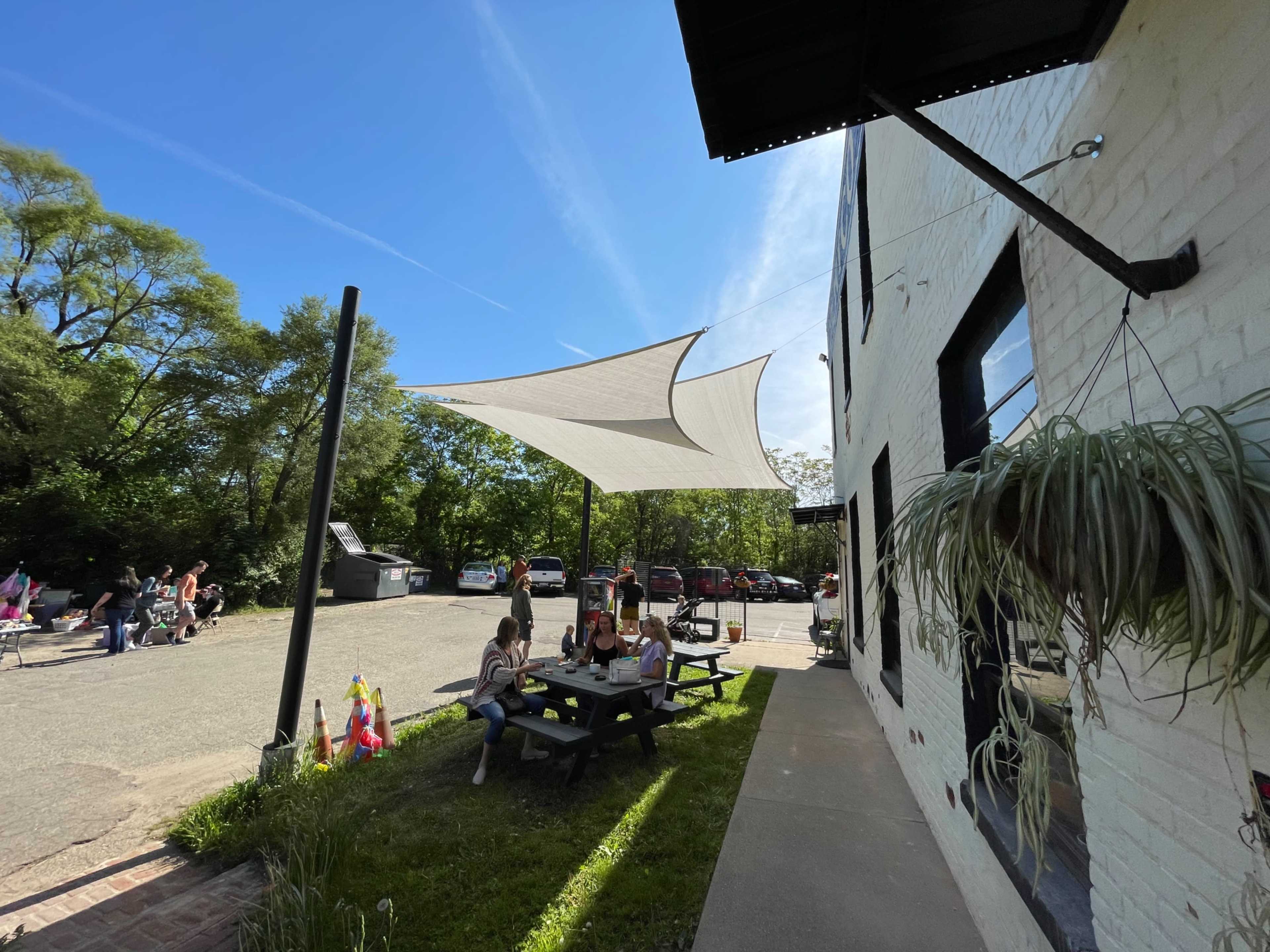 A shaded outdoor seating area with a picnic table is filled with people beside a parking lot surrounded by trees.