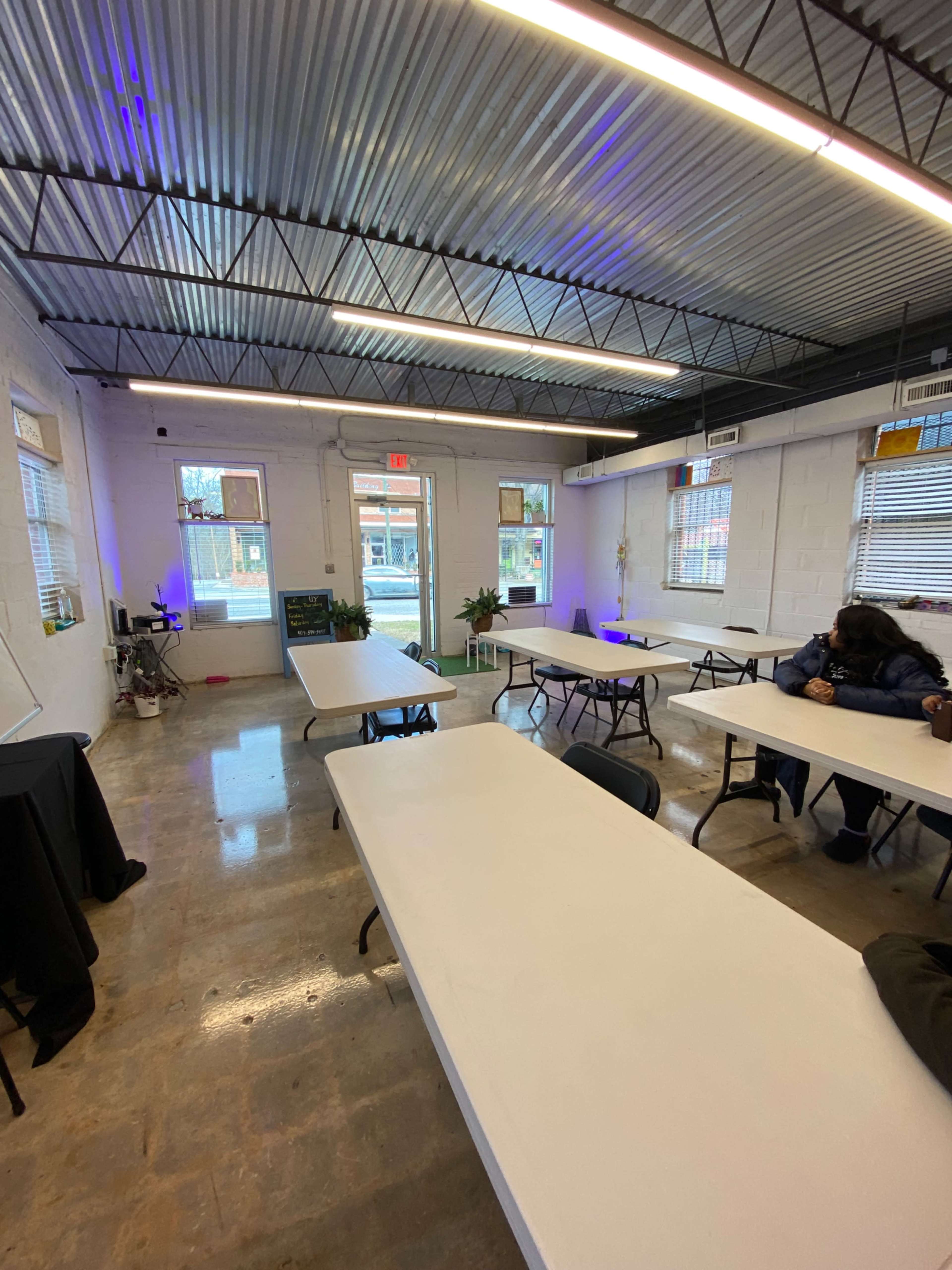 The image shows a bright, sparsely furnished room with several tables arranged in a workshop setup and large windows allowing natural light to enter.