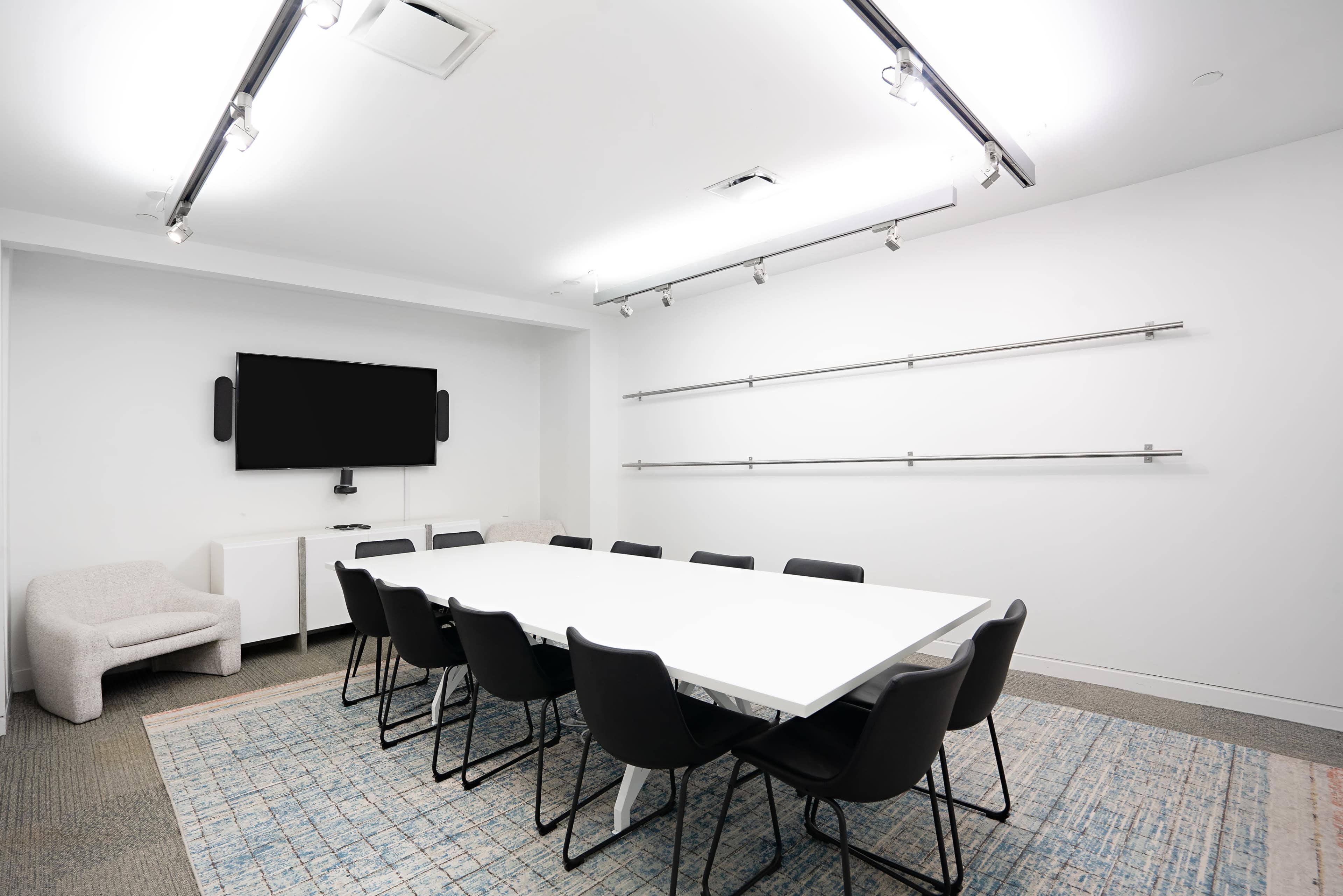 A modern conference room features a long white table surrounded by black chairs, a television mounted on the wall, and minimal decor.