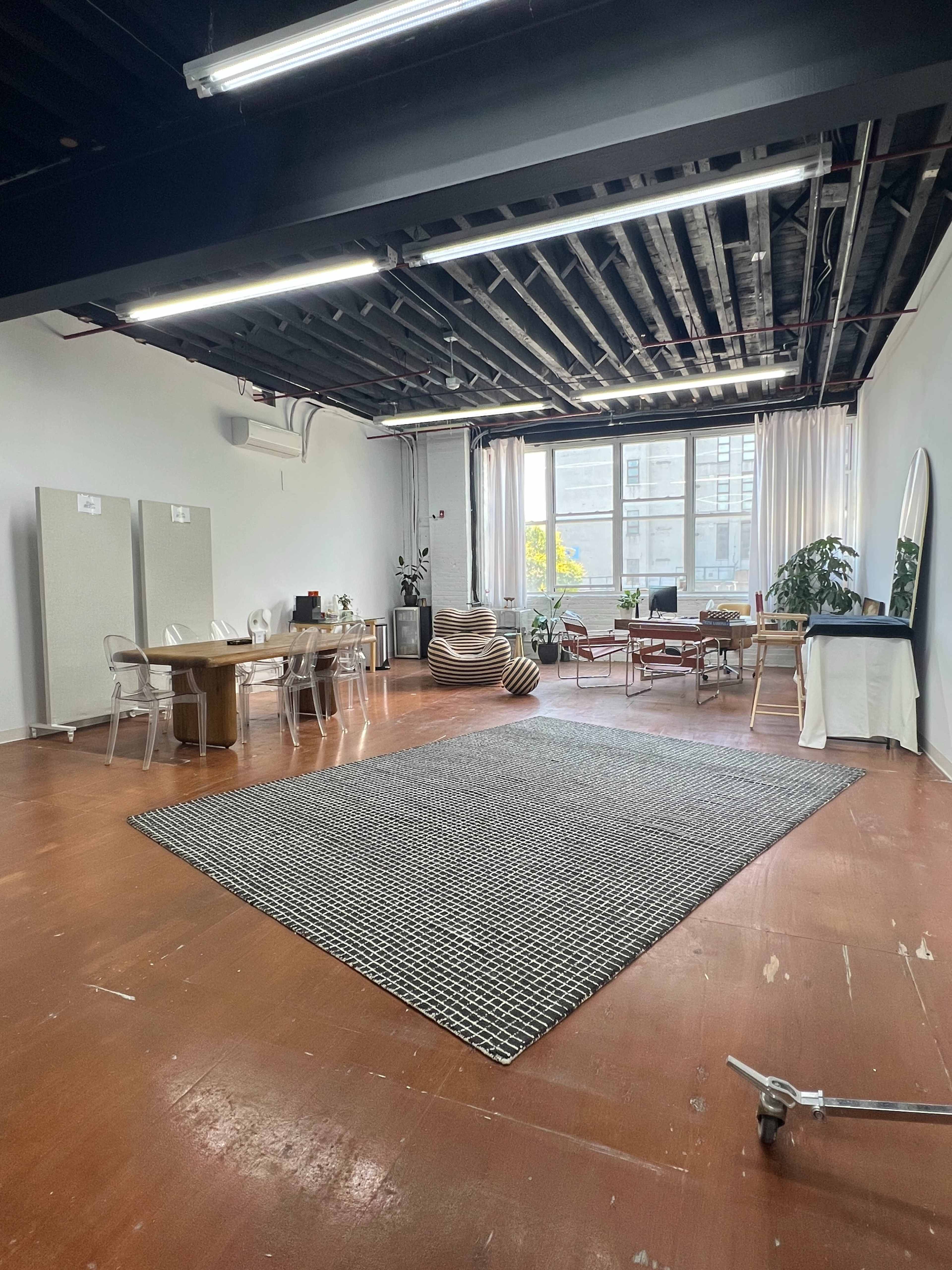 The image shows a spacious, well-lit studio with wooden floors, a large area rug, a dining table, and various seating arrangements.