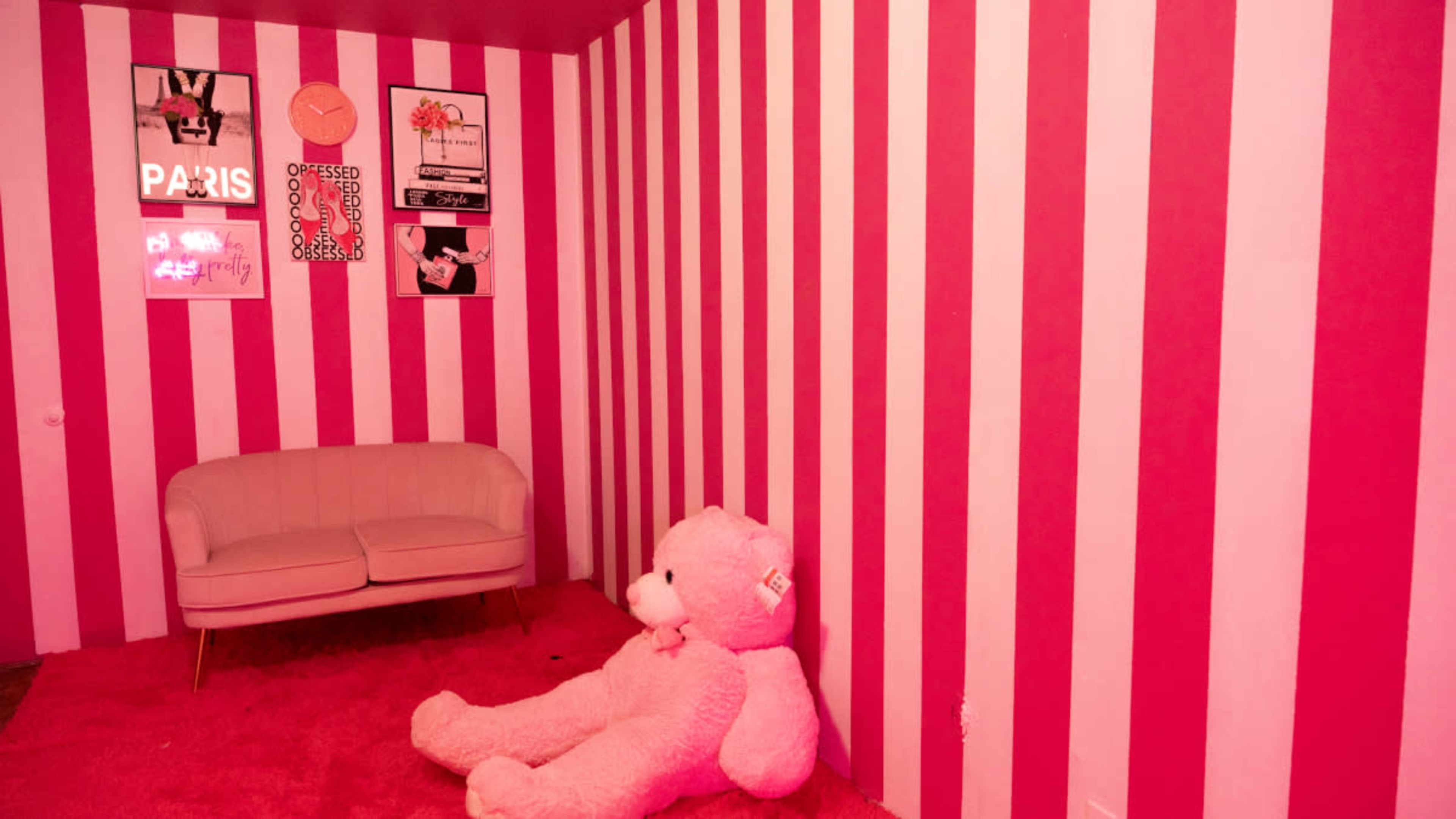 The room features pink and white striped walls, a pink shag rug, a beige couch, and a large pink teddy bear sitting on the floor.