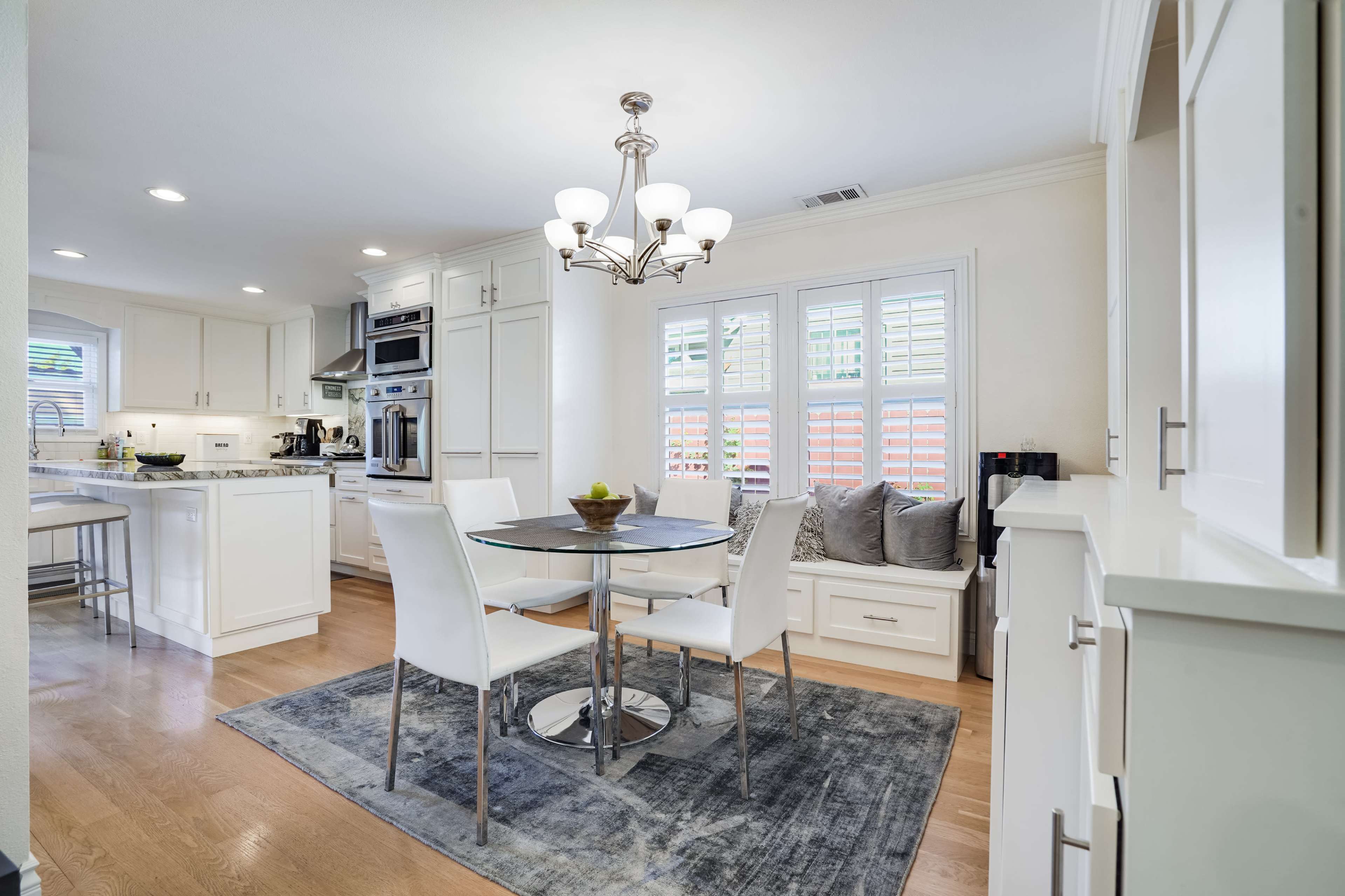A modern kitchen and dining area features a round glass table surrounded by white chairs, with a chandelier above and a window displaying plants outside.
