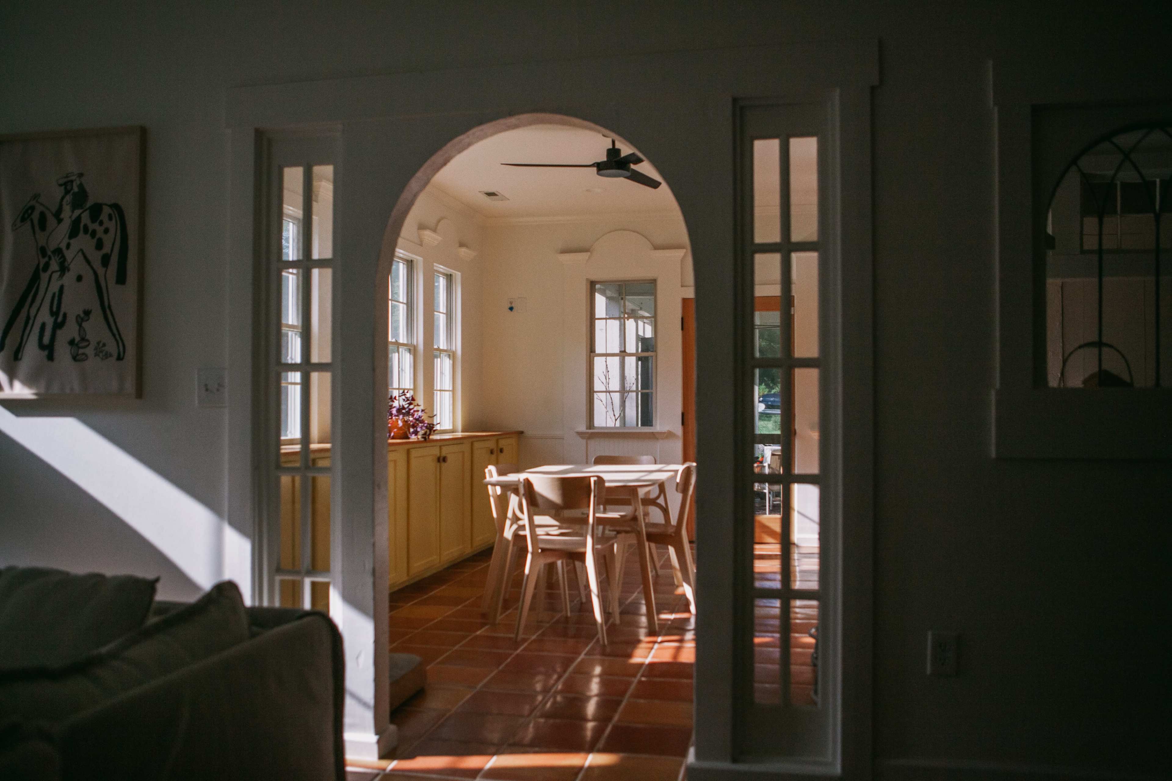 A sunlit room is visible through an arched doorway, featuring a wooden table surrounded by chairs and large windows that let in natural light.