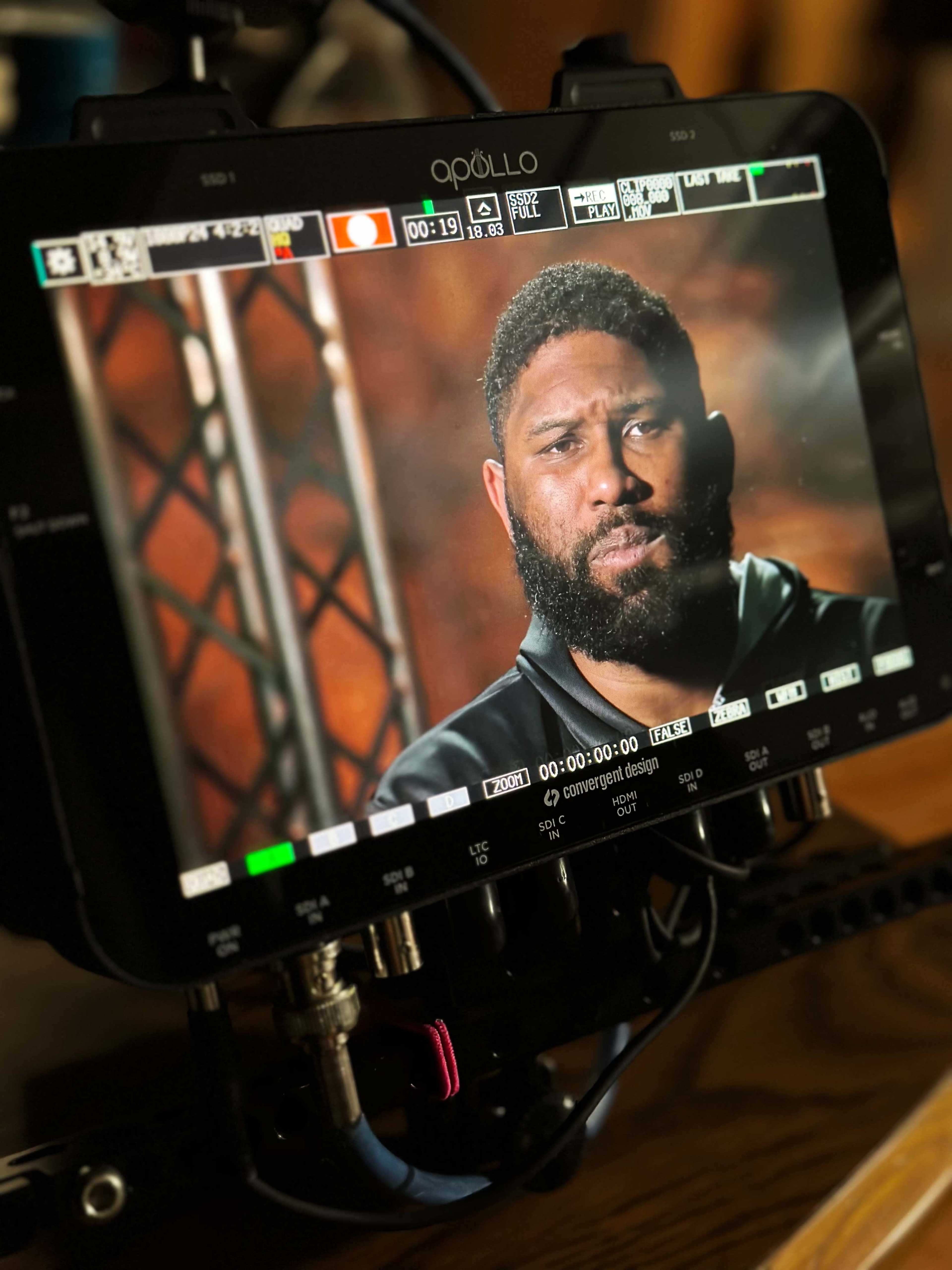 The image shows a digital monitor displaying a close-up of a man with a beard during a video shoot, with the user interface visible on the screen.