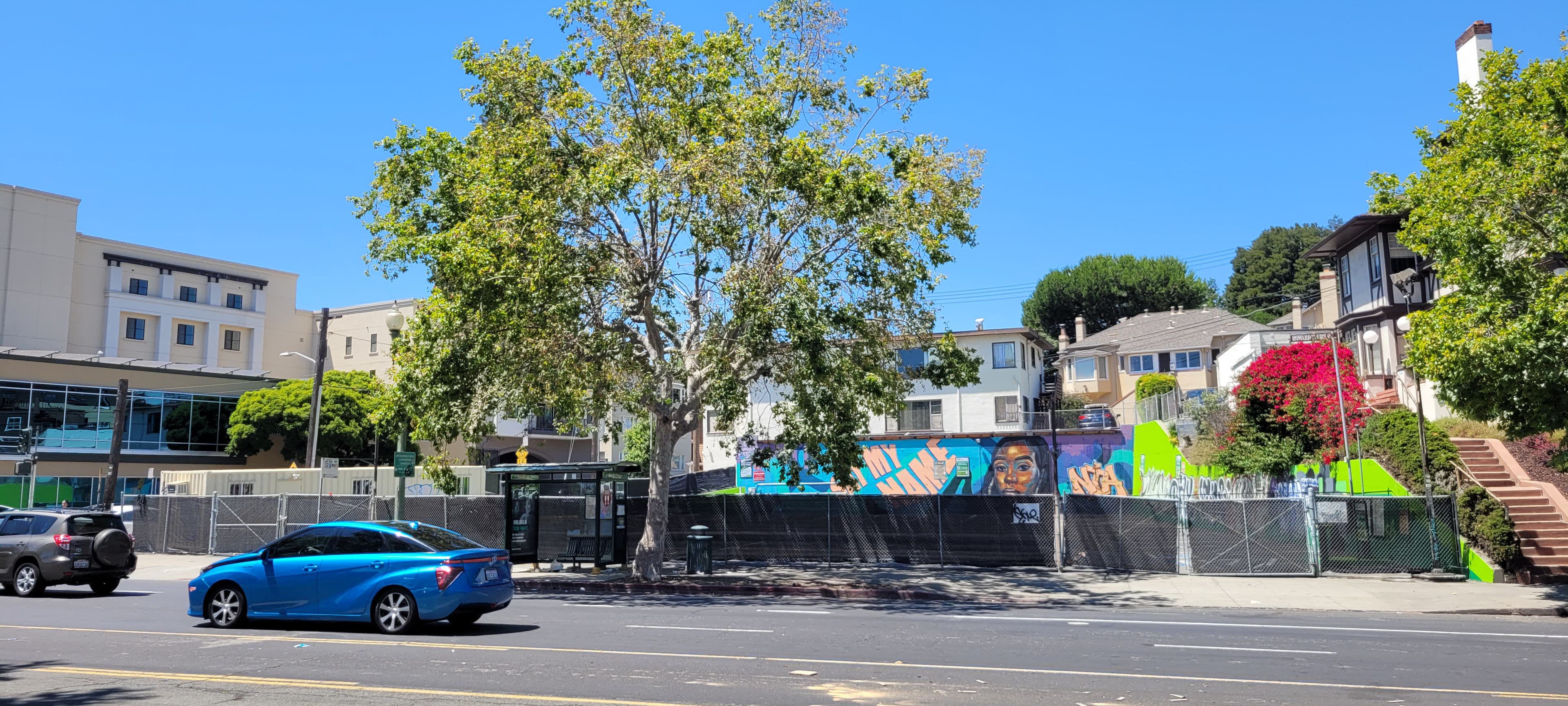 A blue car is parked on the street in front of a fenced area featuring a vibrant mural and a large tree, with residential buildings in the background.