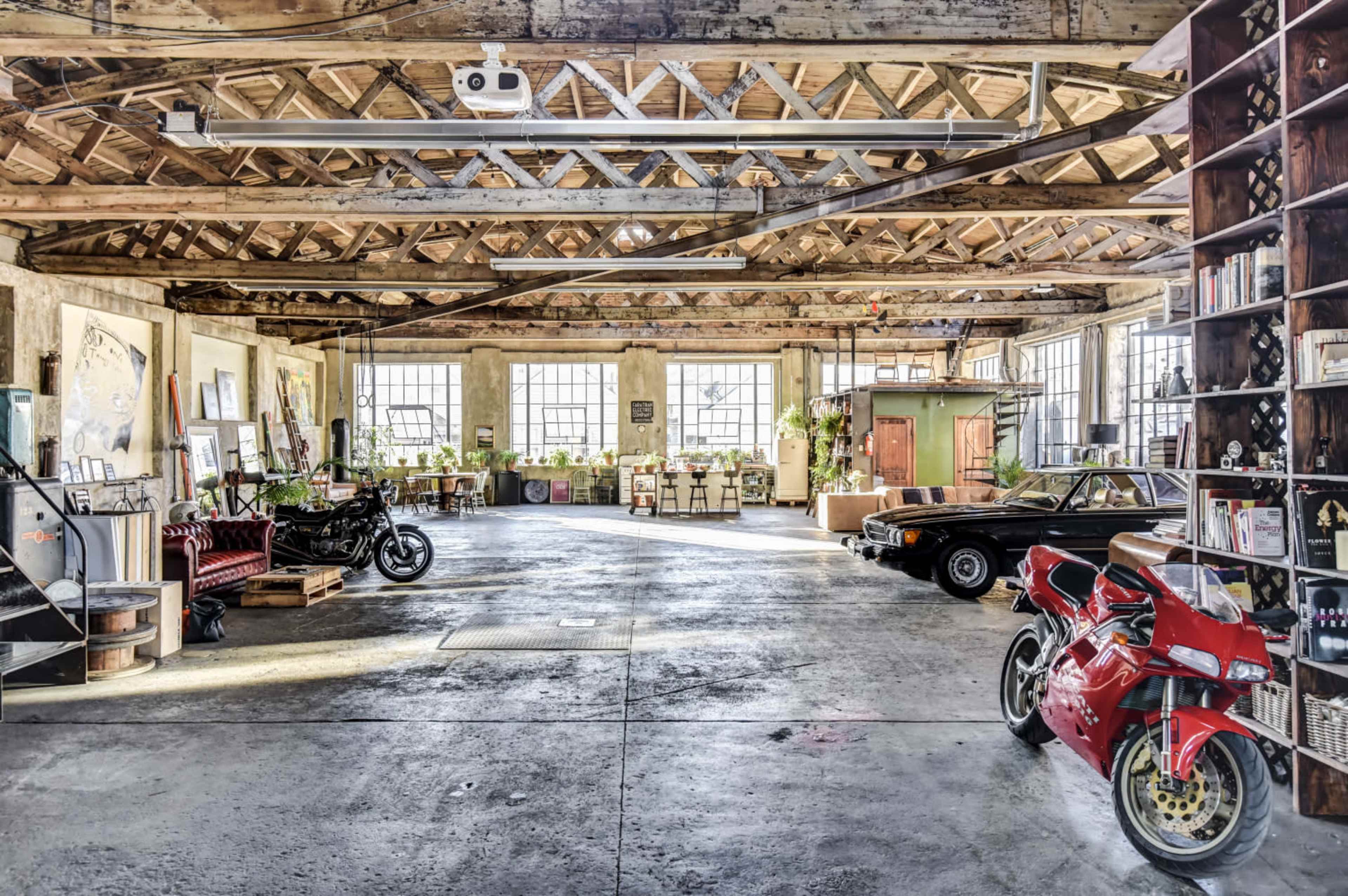 A large, open industrial space featuring vintage cars, motorcycles, and various seating arrangements under a wooden-beamed ceiling.