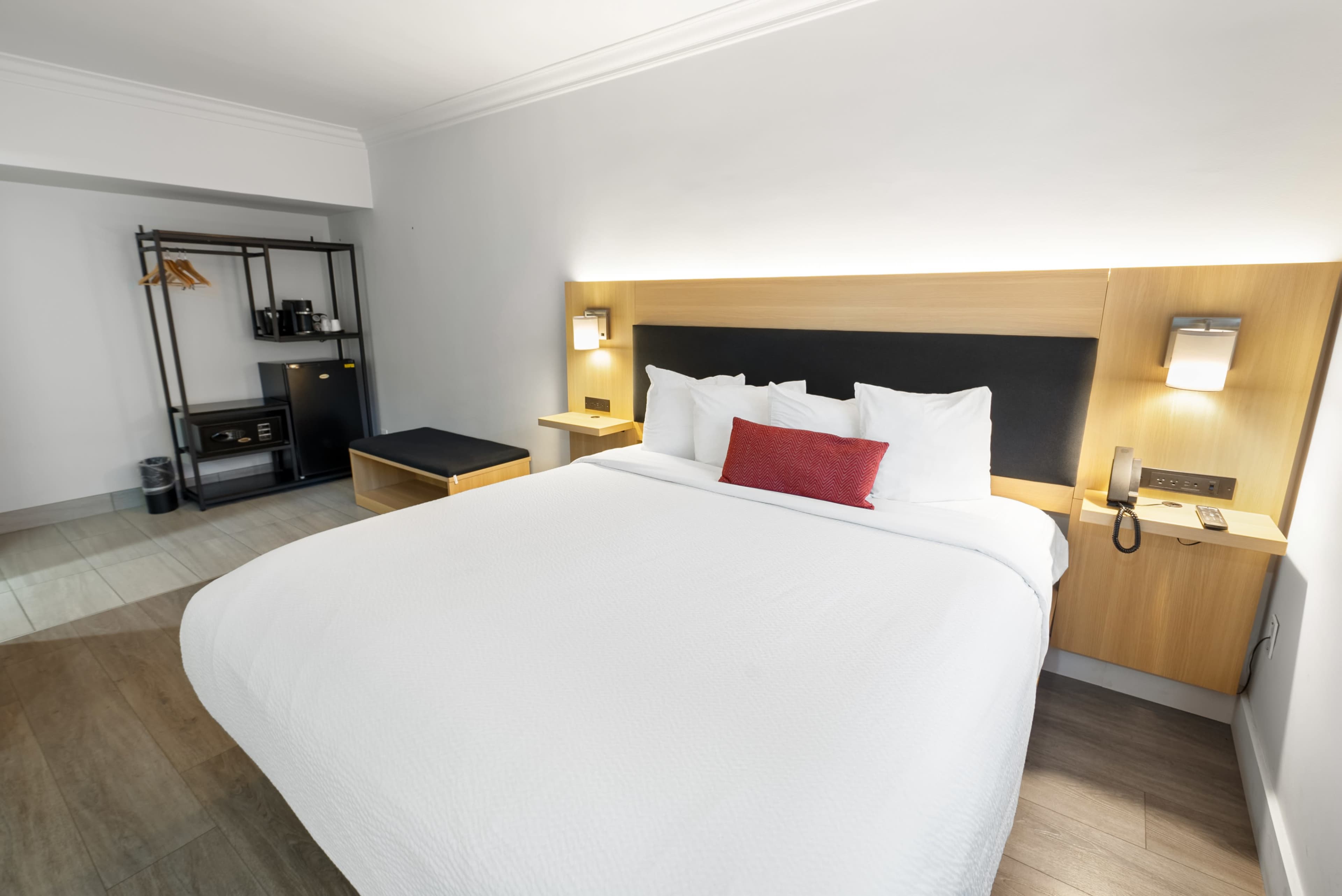 A modern hotel room featuring a large bed with white linens, a red accent pillow, and a wooden headboard, along with a wardrobe, seating area, and minibar.