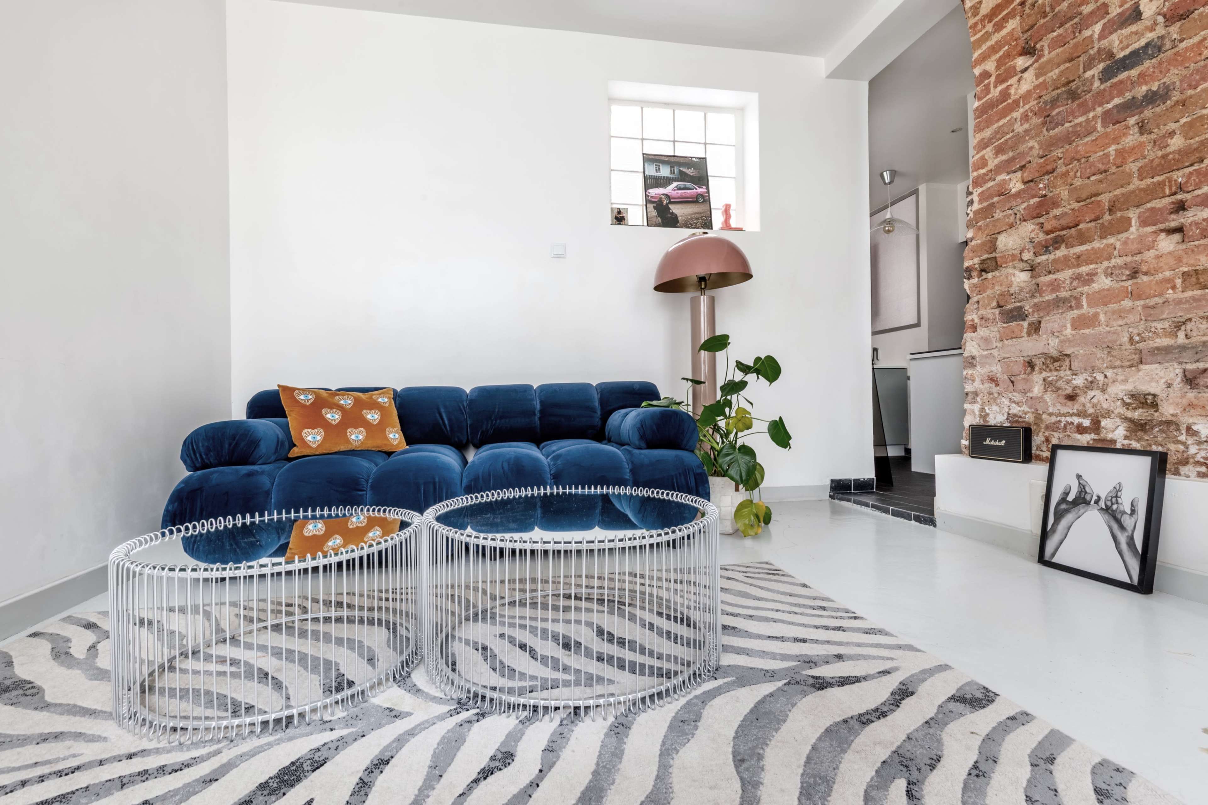 The image shows a modern living room featuring a blue velvet sofa, two white metal coffee tables, and a zebra-patterned rug, with exposed brick walls and a floor lamp in the background.