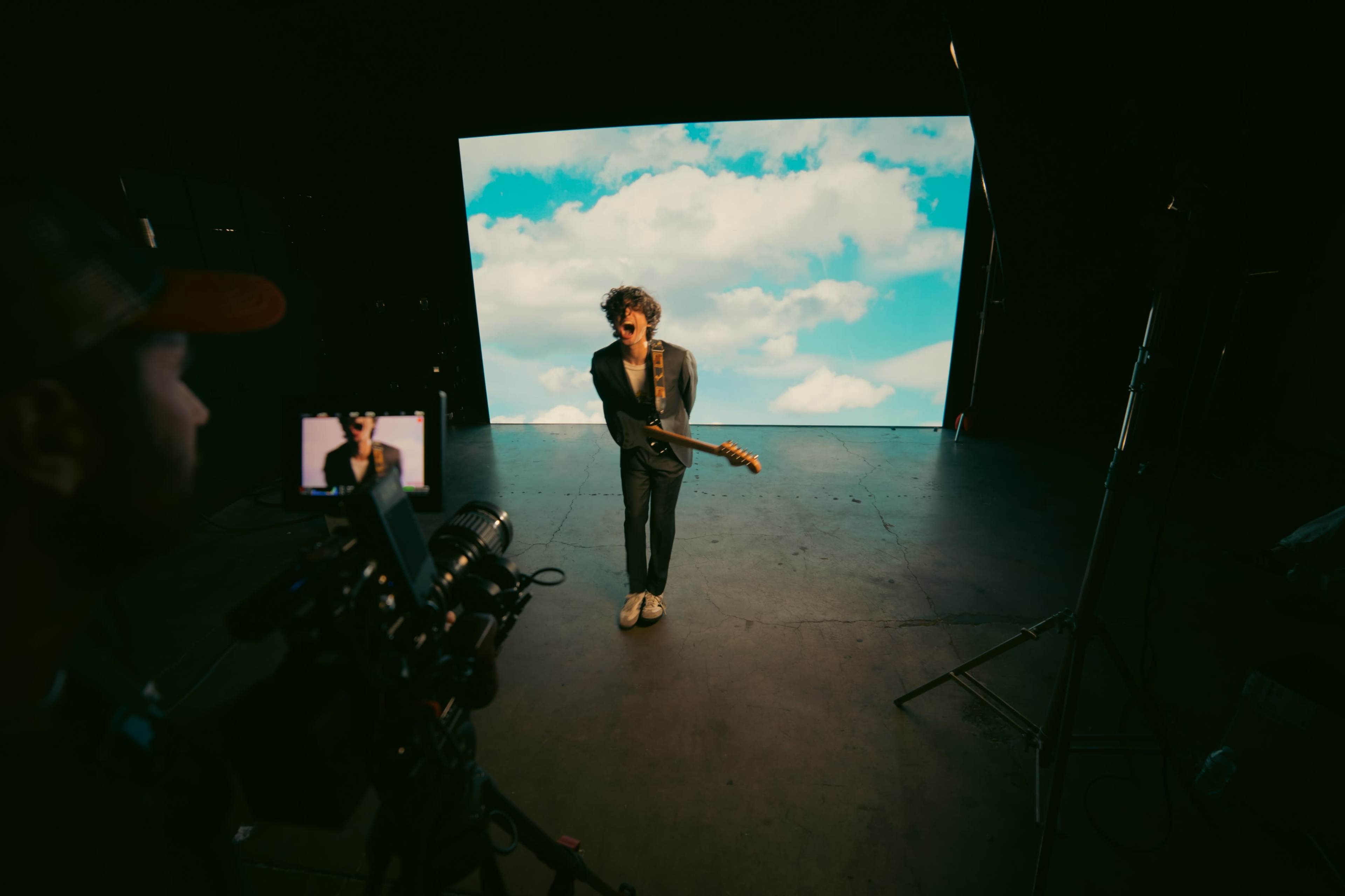 A musician stands on stage with a guitar in front of a large screen displaying a blue sky with clouds, while a camera operator captures the performance.