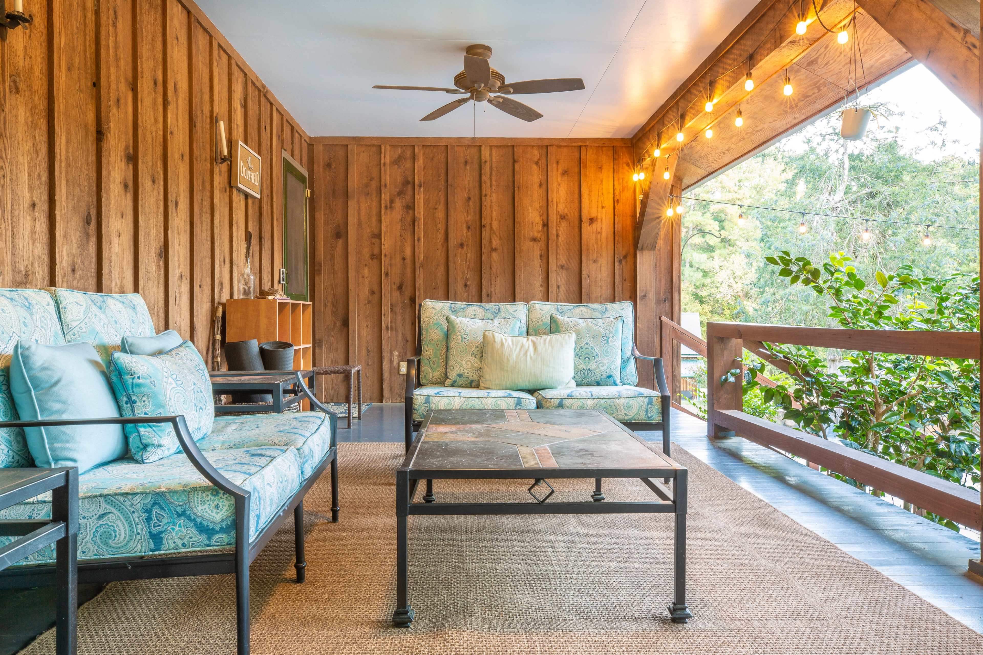 The image shows a cozy porch with wooden walls, a ceiling fan, two upholstered sofas, and a coffee table, illuminated by hanging string lights.