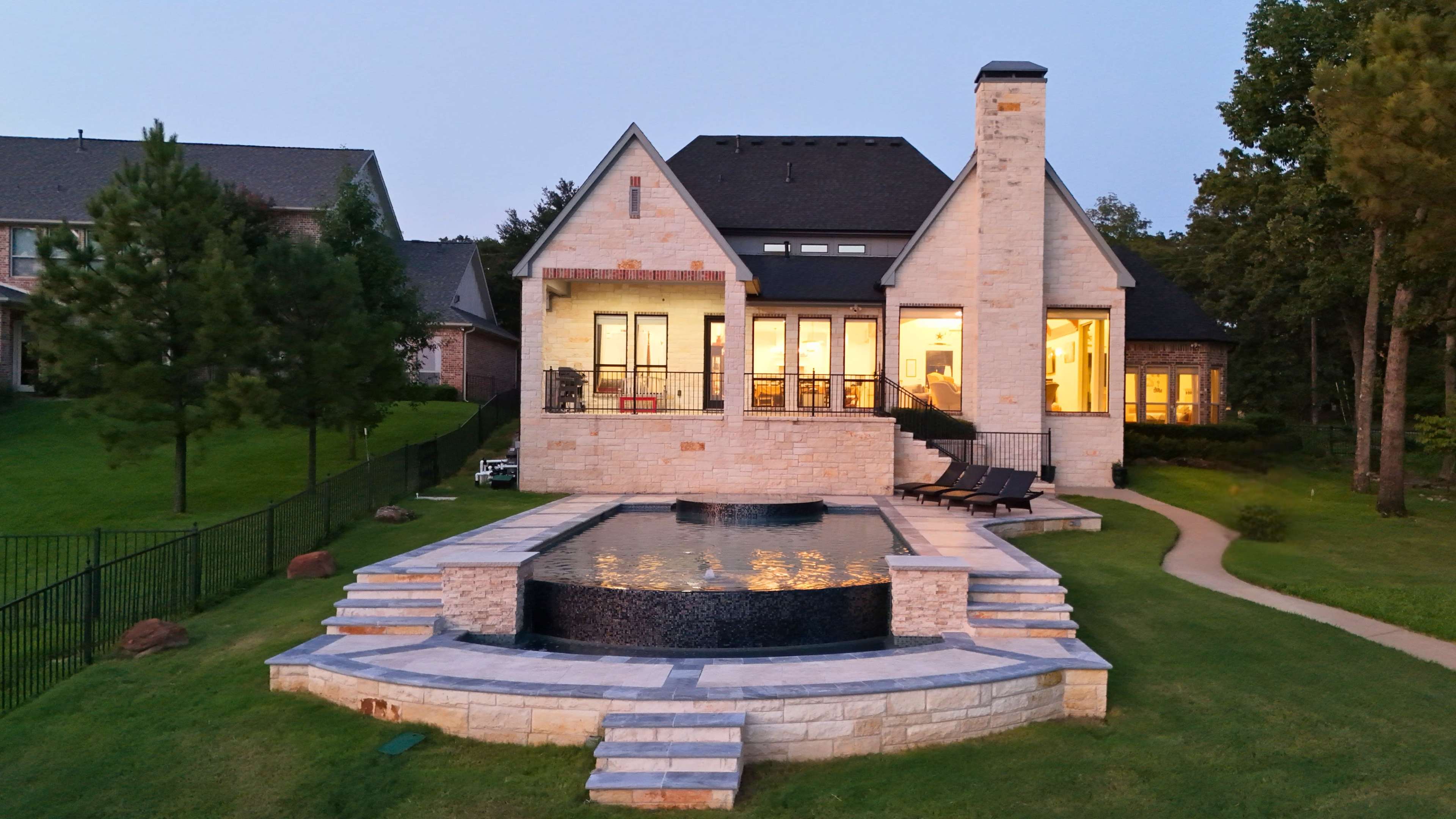 The image shows a modern two-story stone house with a swimming pool and patio area, set on a grassy landscape with trees and neighboring homes in the background.