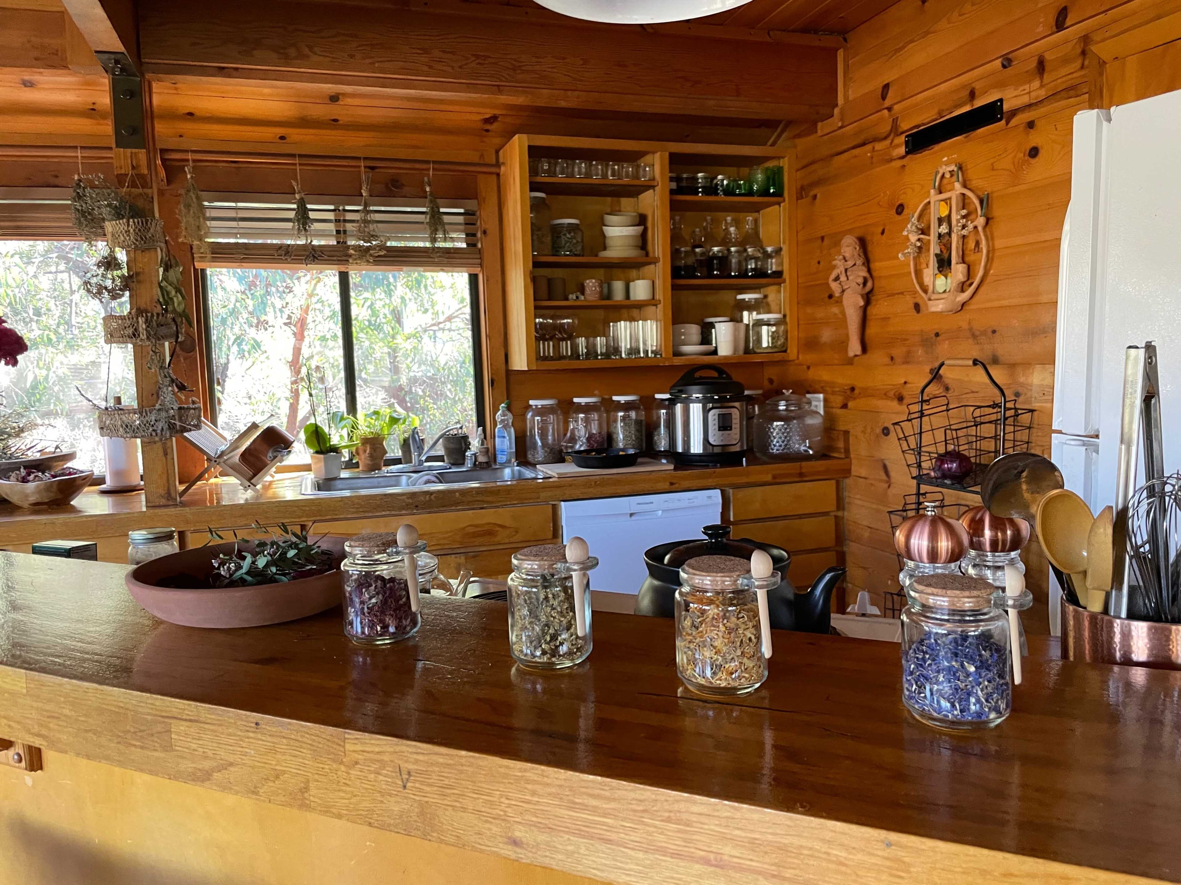 A wooden kitchen features jars of herbs lined up on a counter, with shelves of glassware and cooking utensils visible in the background.