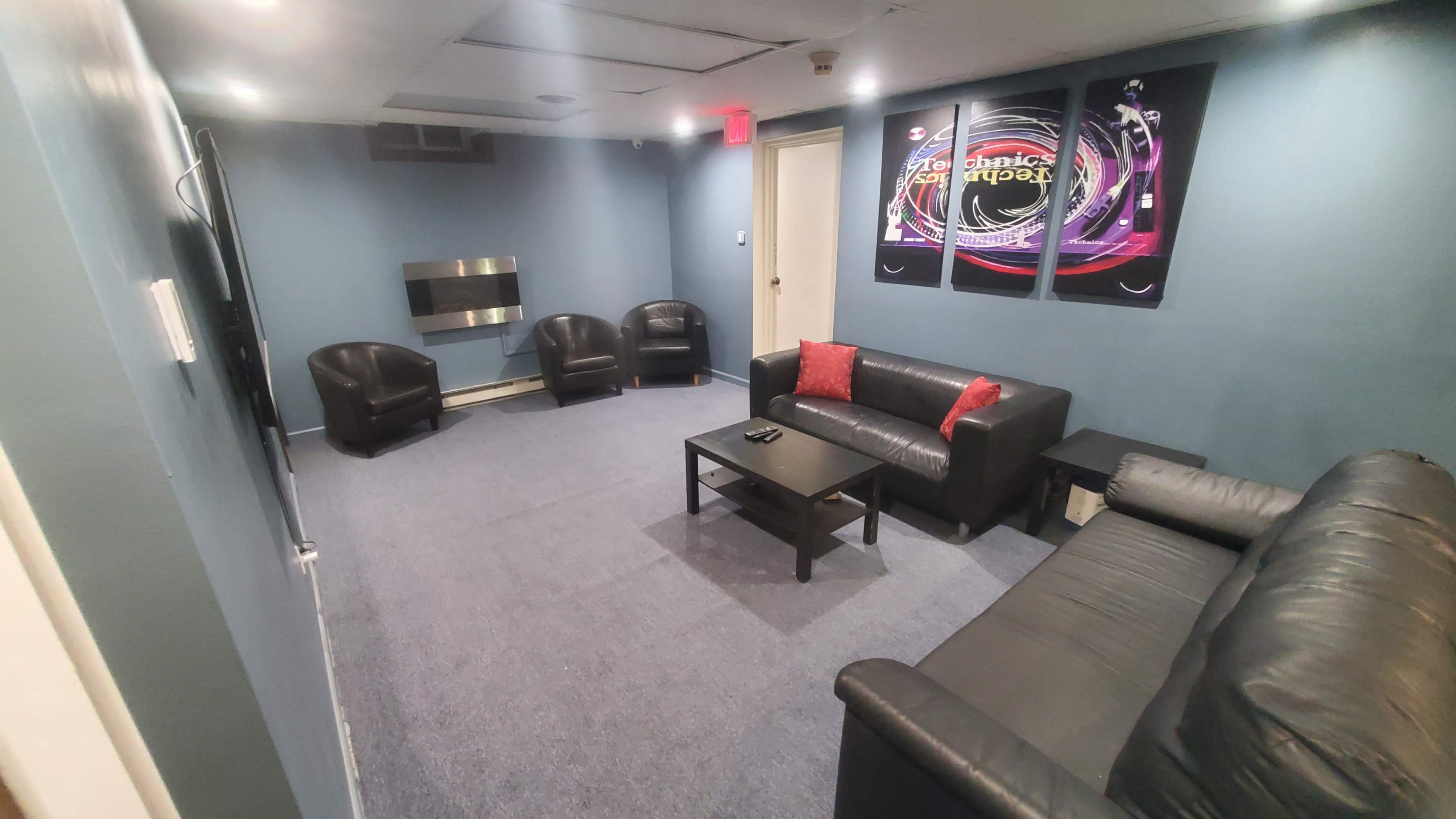 A small lounge area with two black leather sofas, two chairs, a coffee table, and wall art, all arranged around a television and a wall-mounted electric fireplace.