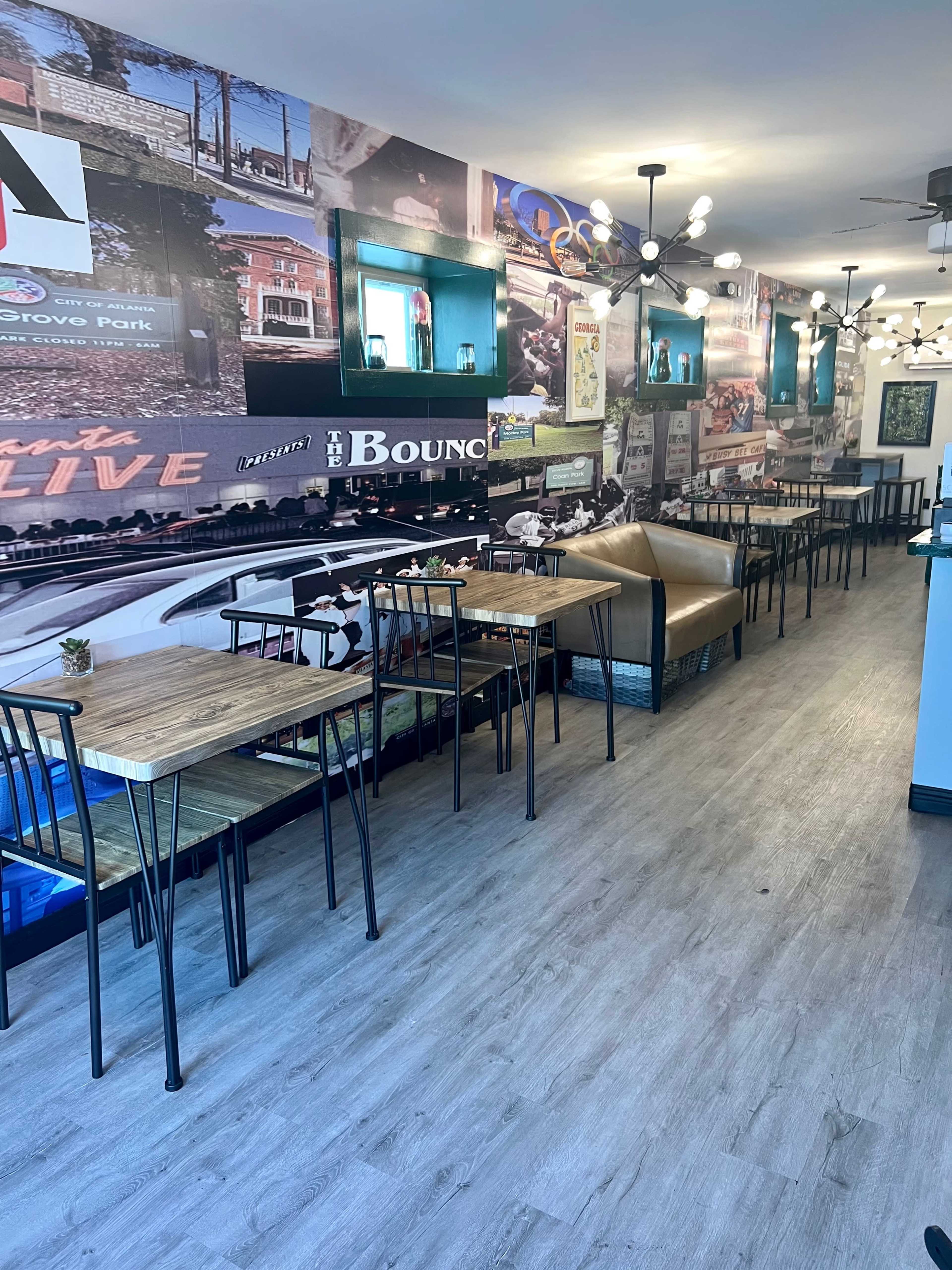 A long, narrow restaurant or café interior with tables and chairs arranged along one side and a decorative wall featuring various vintage-style advertisements.