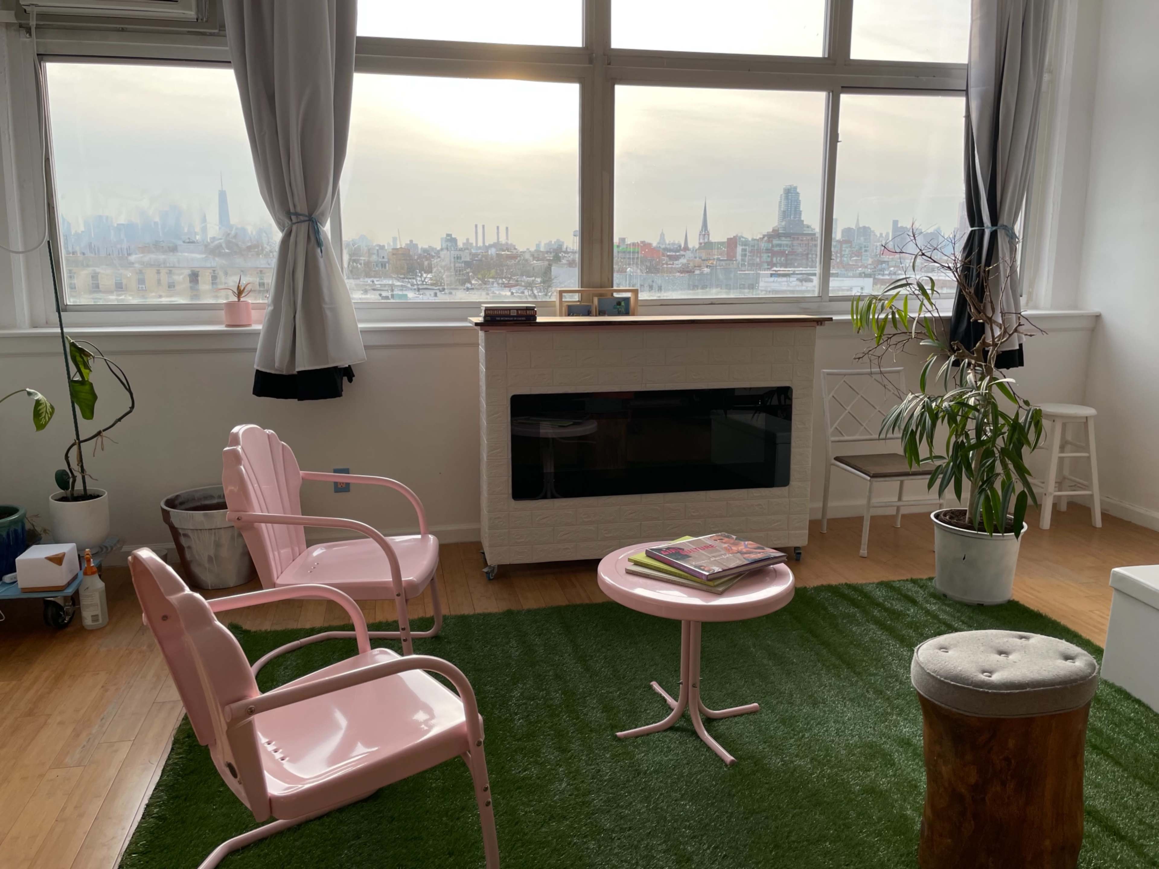 A bright room features pink chairs and a small table on a green rug, with large windows showcasing a city skyline in the background.