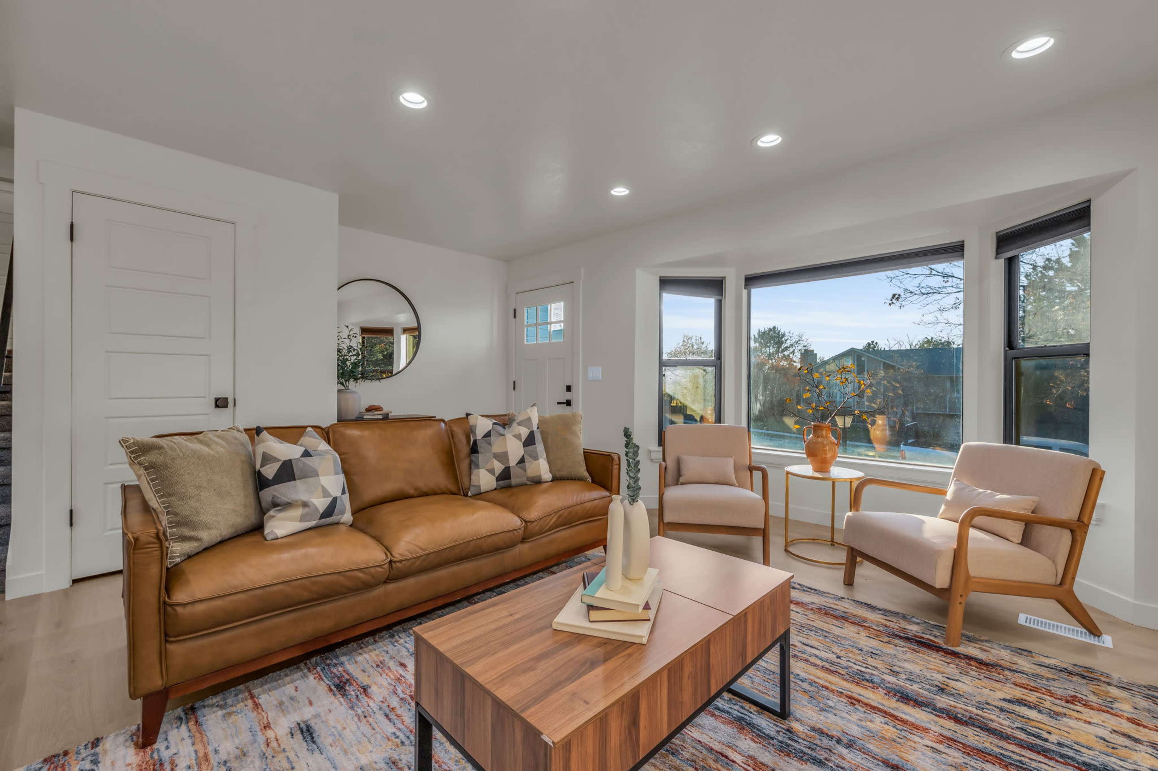 A well-lit living room features a brown leather sofa, two armchairs, a coffee table, and large windows with a view of the outdoors.