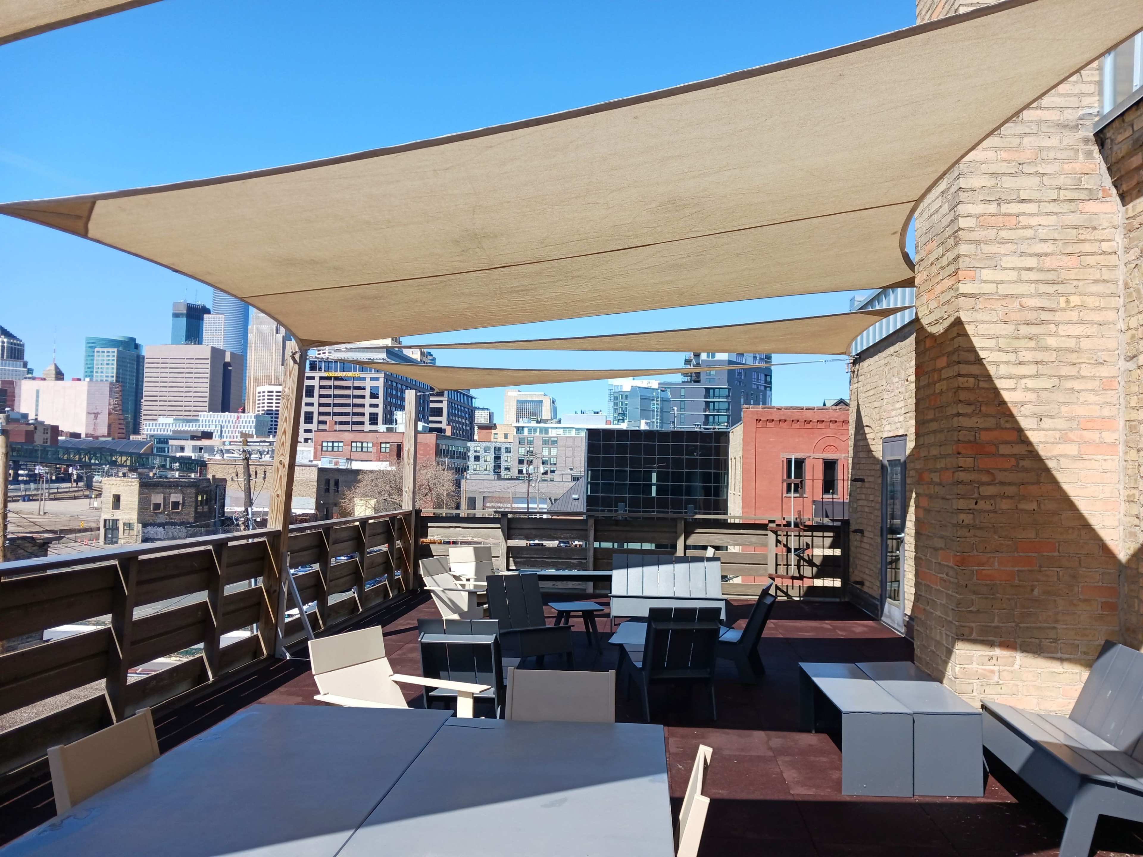 The image shows a rooftop terrace with several tables and chairs shaded by large fabric canopies, overlooking a city skyline.