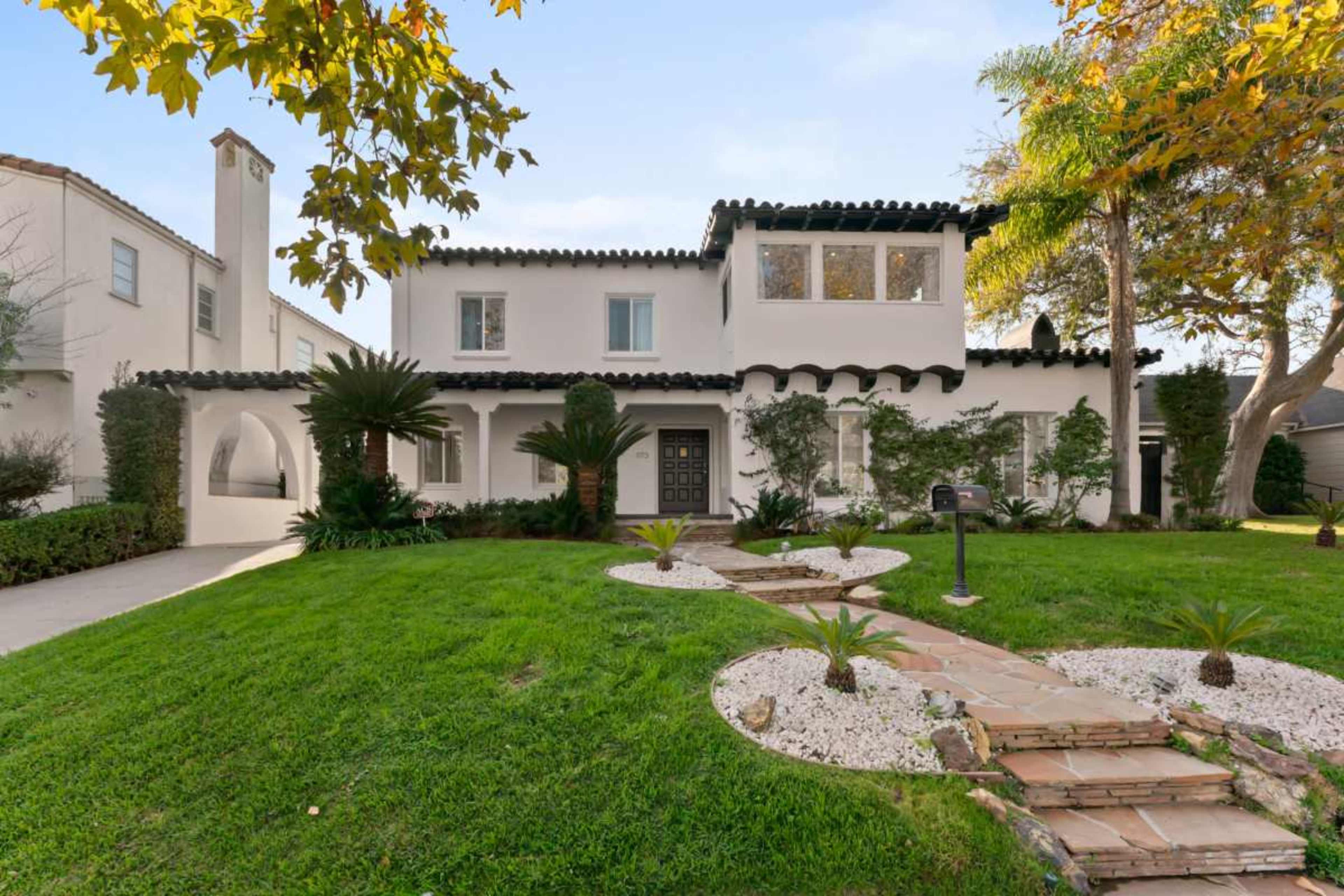 A two-story white house with a tiled roof and green landscaping features a pathway lined with decorative stones and plants.