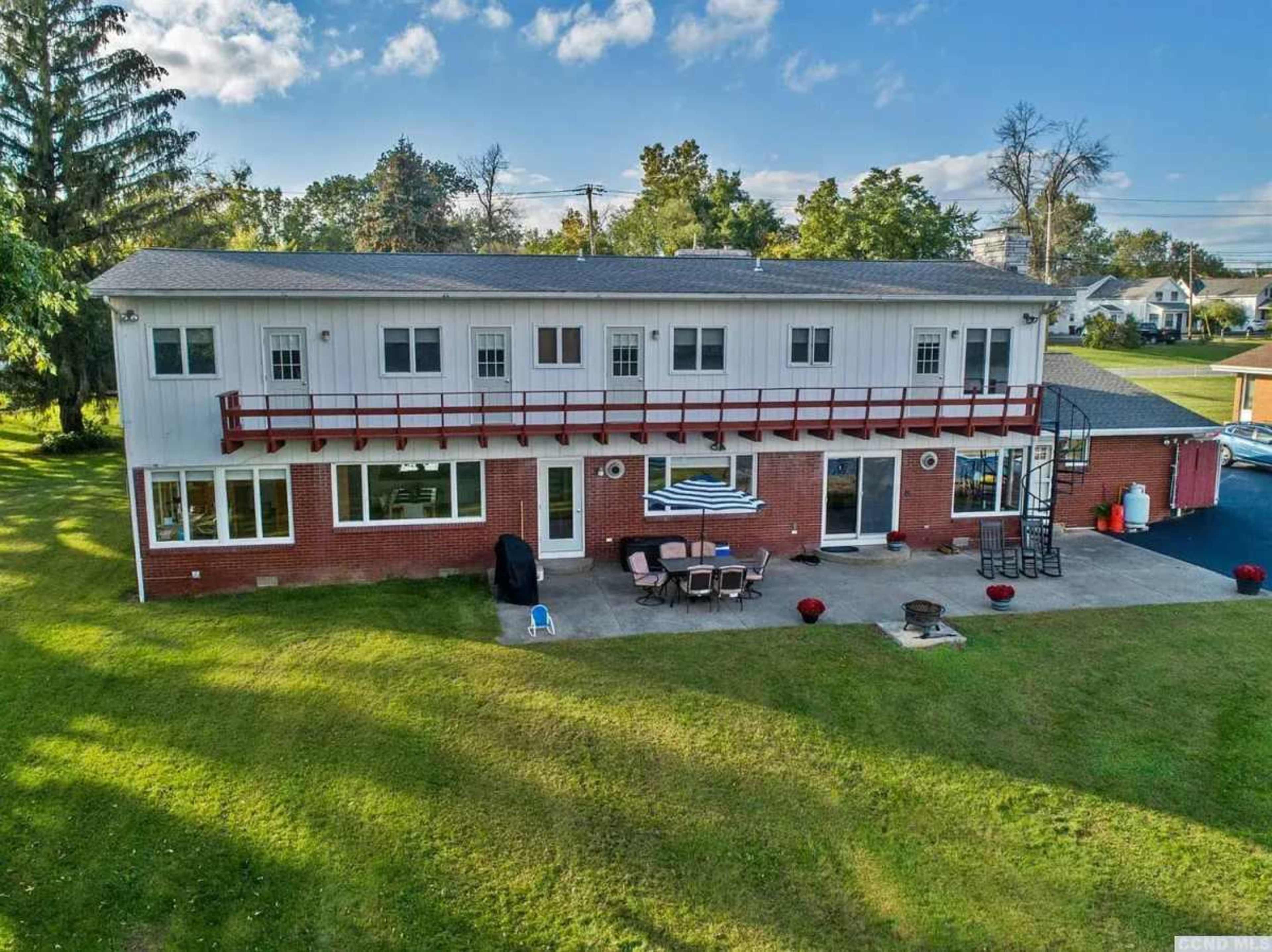 The image shows a two-story house with a brick and siding exterior, featuring a patio and outdoor seating area in the backyard.