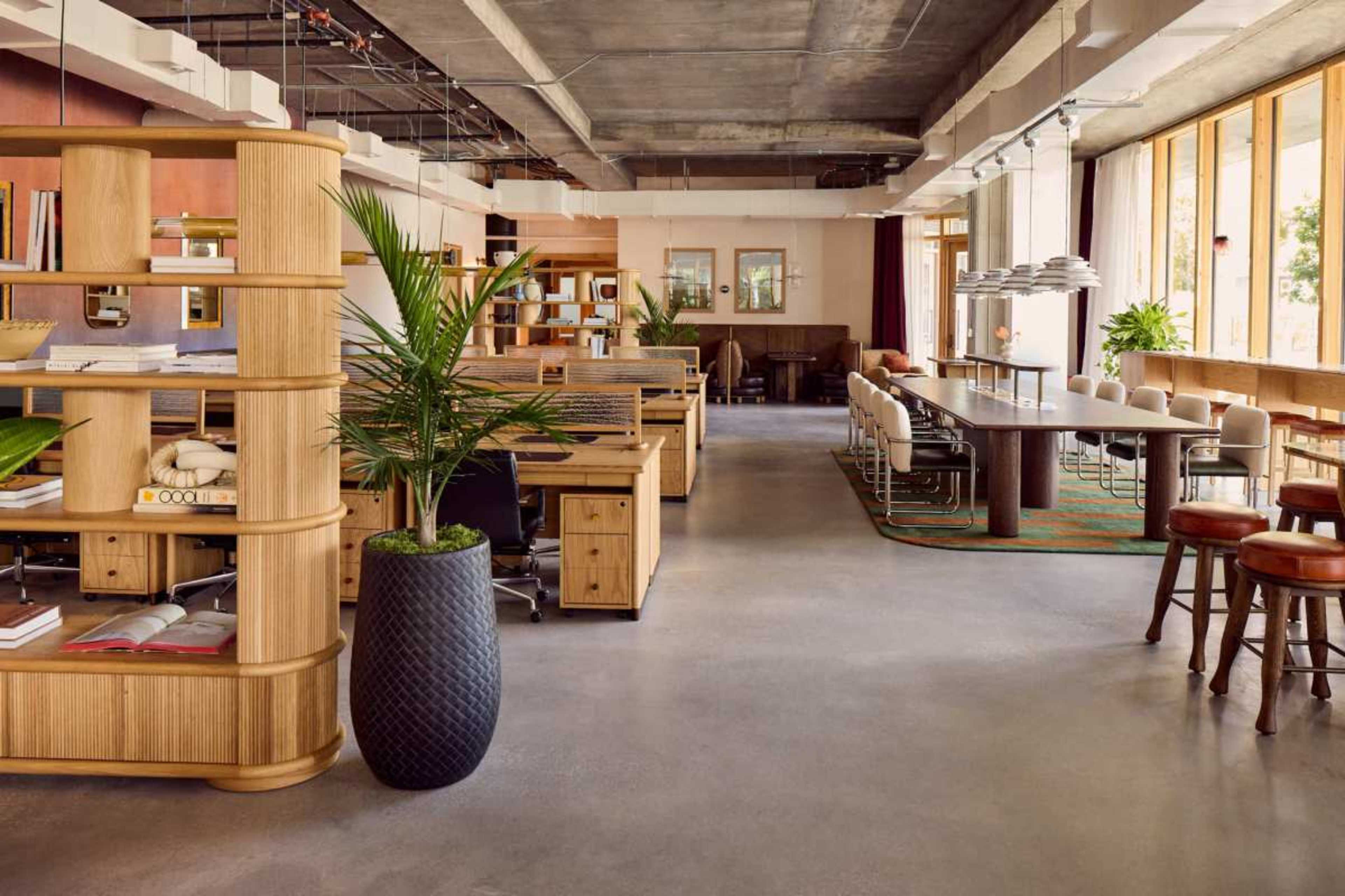 The image shows a modern office space featuring wooden furniture, a large communal table, and plants, all under an industrial-style ceiling with large windows.