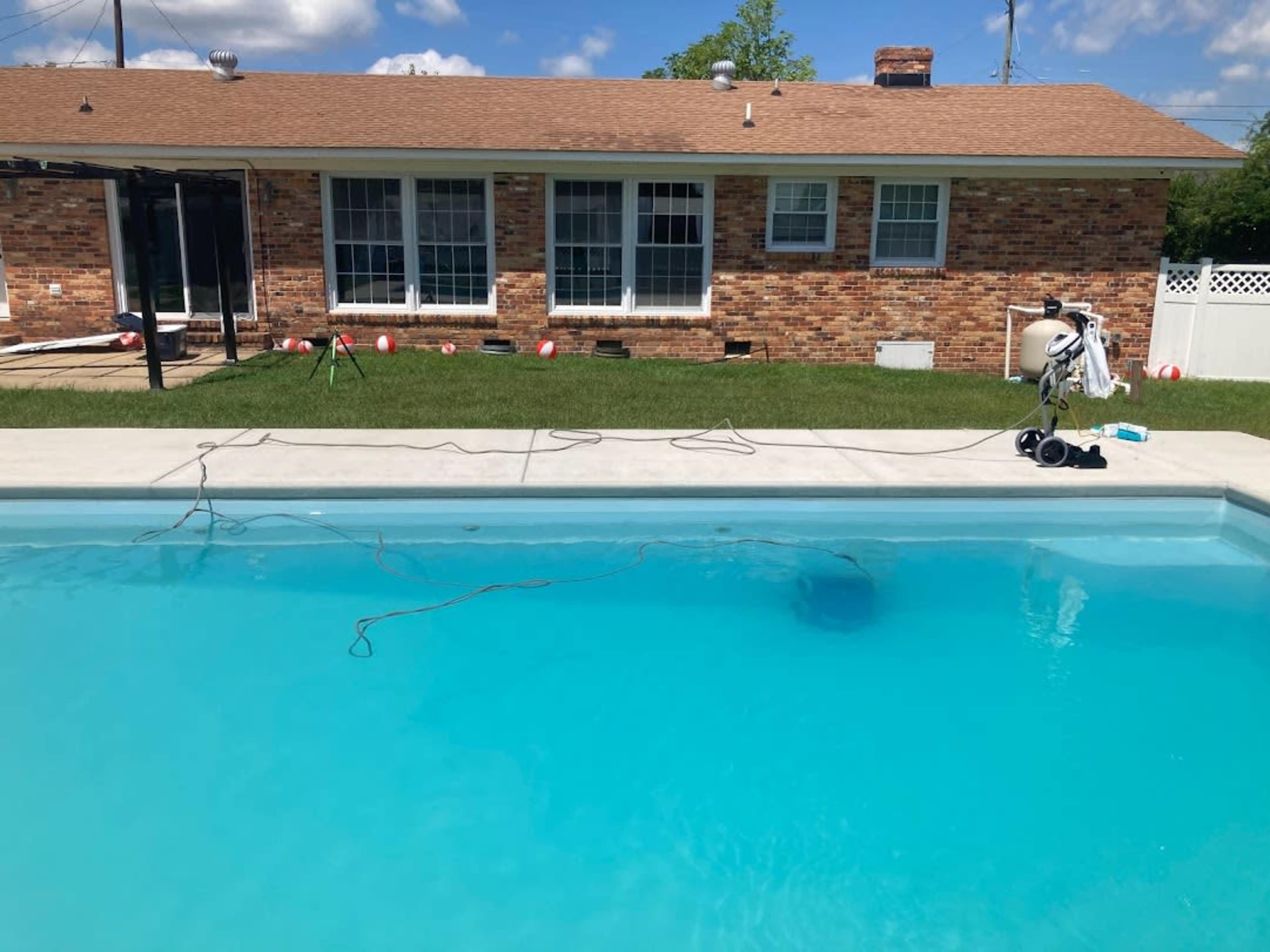 A swimming pool with clear blue water is set in a backyard, alongside a brick house with large windows and a grass lawn.