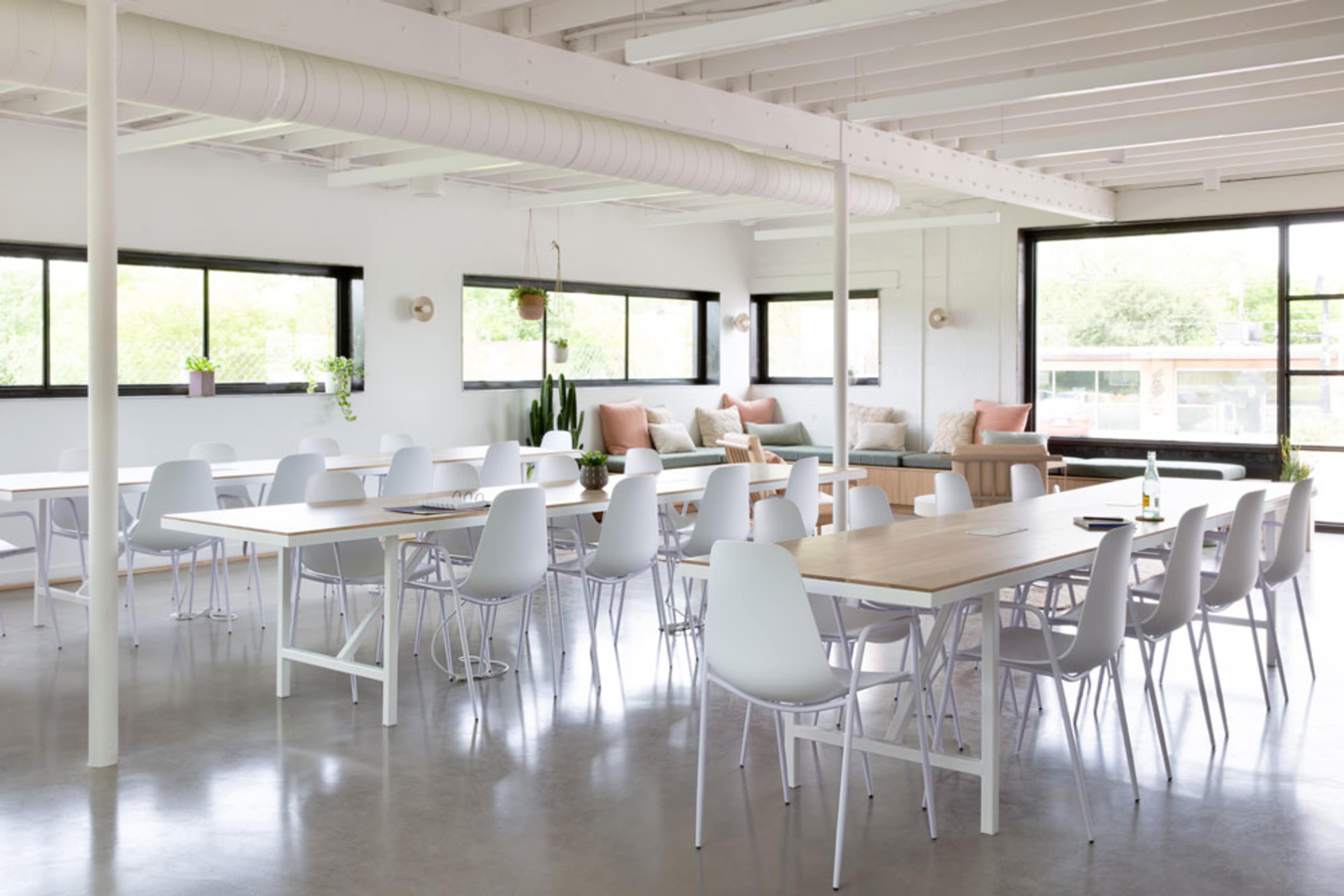 A modern, open-concept workspace with white tables, chairs, and large windows, featuring a comfortable seating area along one wall.