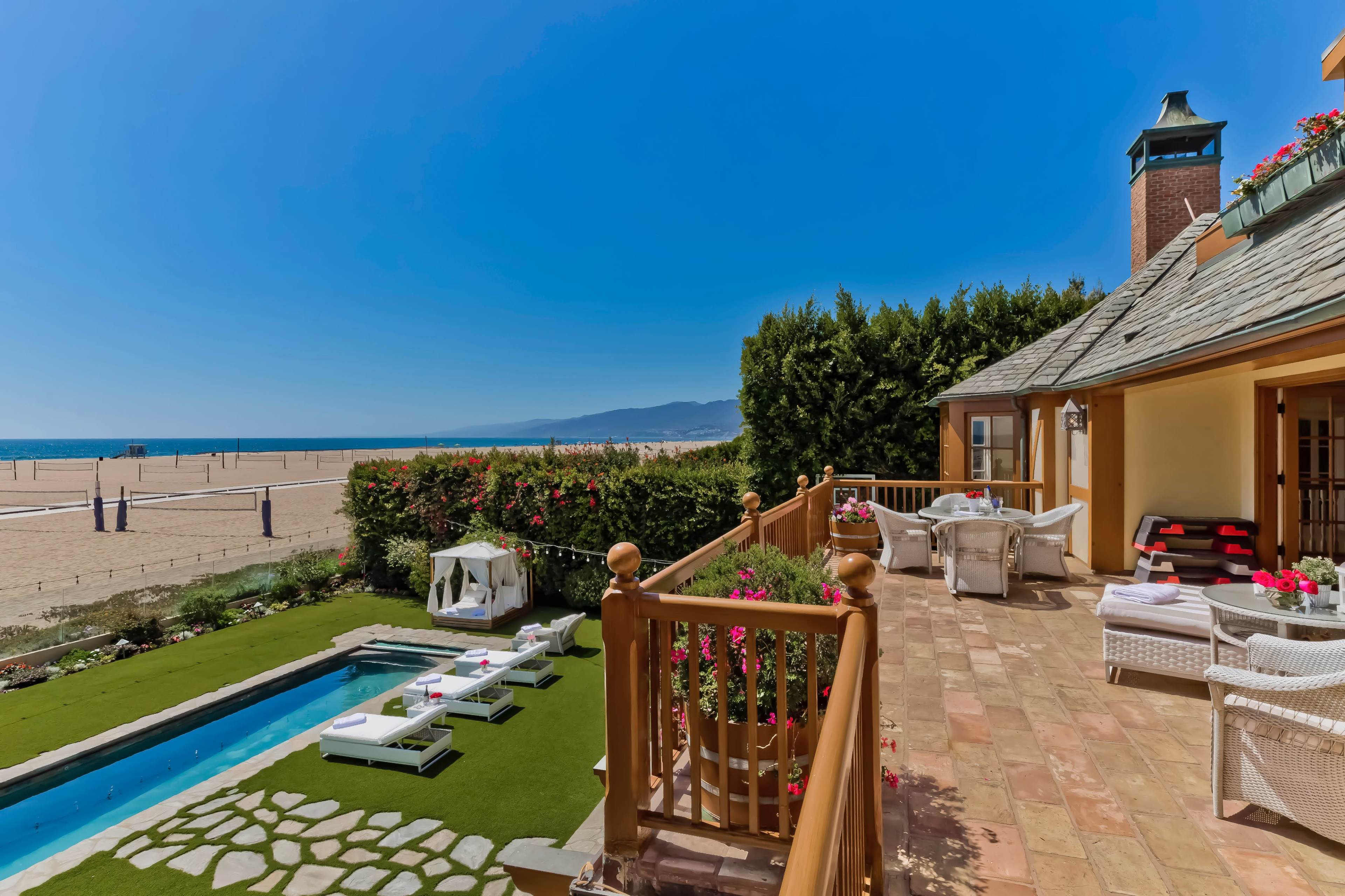 The image shows a beachside patio with a swimming pool, lounge chairs, and a view of the ocean and sandy beach in the background.