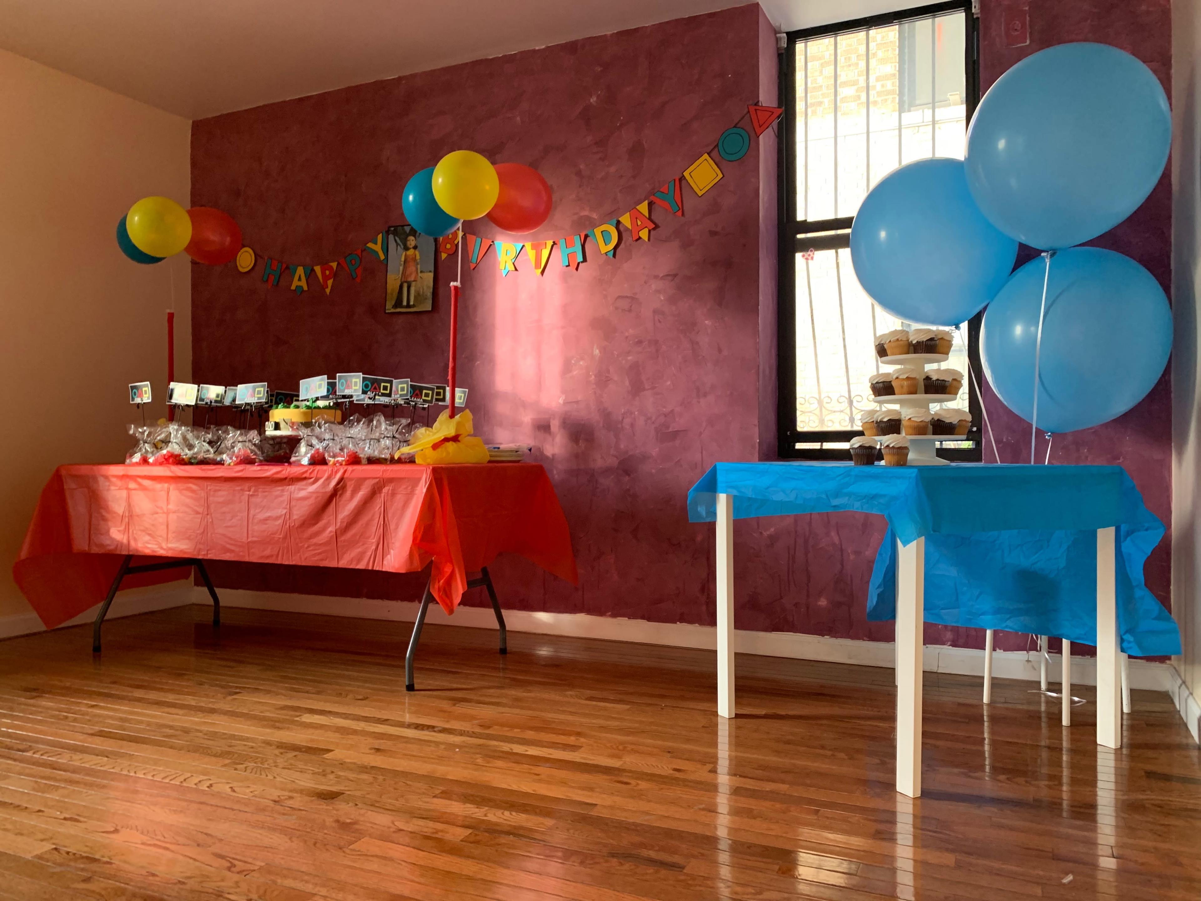The scene features a room decorated for a birthday party, with colorful balloons and two tables set up with treats and decorations against a pink wall.