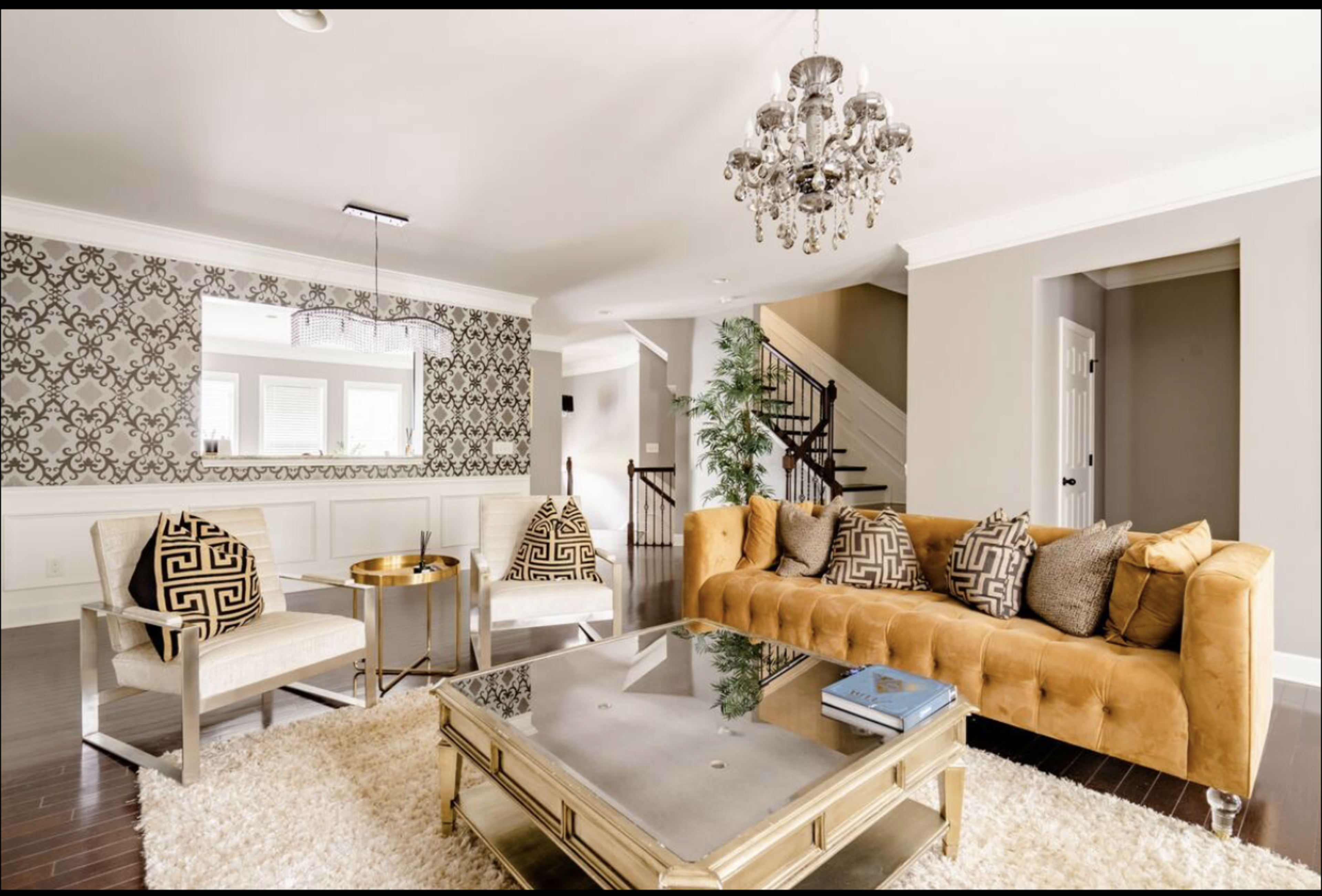 A well-furnished living room features a tan tufted sofa, two patterned armchairs, a glass coffee table, and a chandelier, with decorative wallpaper and hardwood flooring.