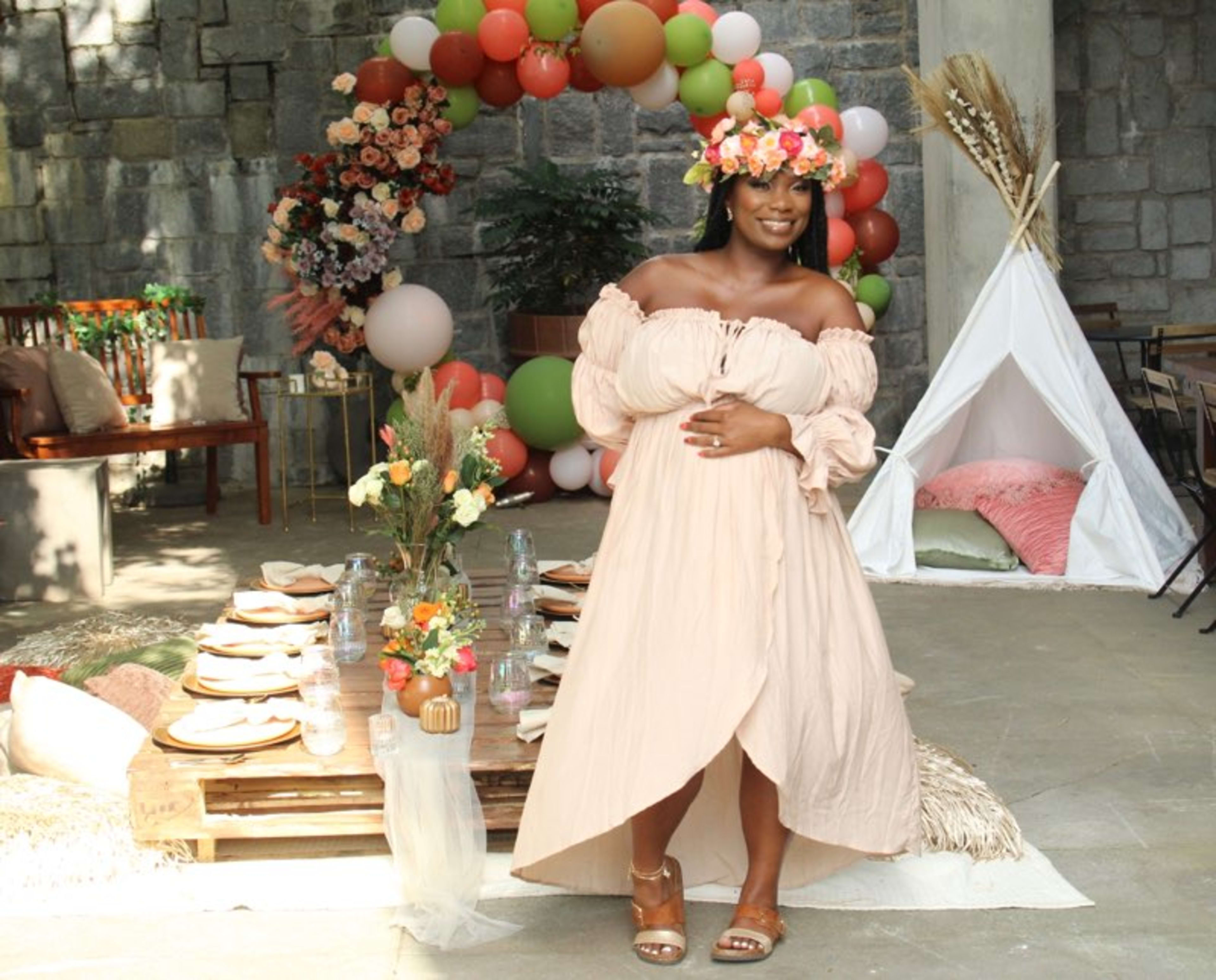 Une femme debout devant une table en extérieur avec des ballons boho pour une baby shower neutre.