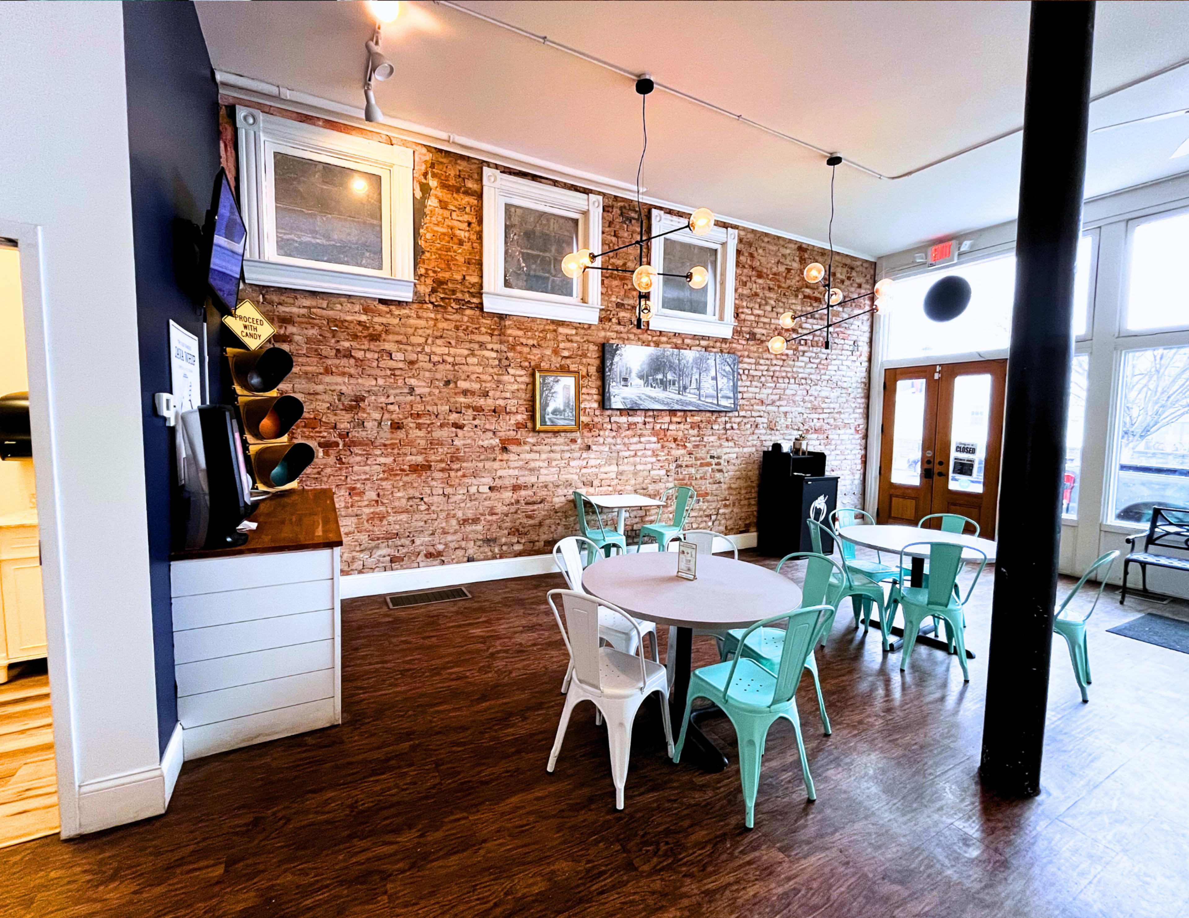 The image shows a cozy café interior featuring wooden floors, exposed brick walls, and a mix of green and white furniture, including tables and chairs.