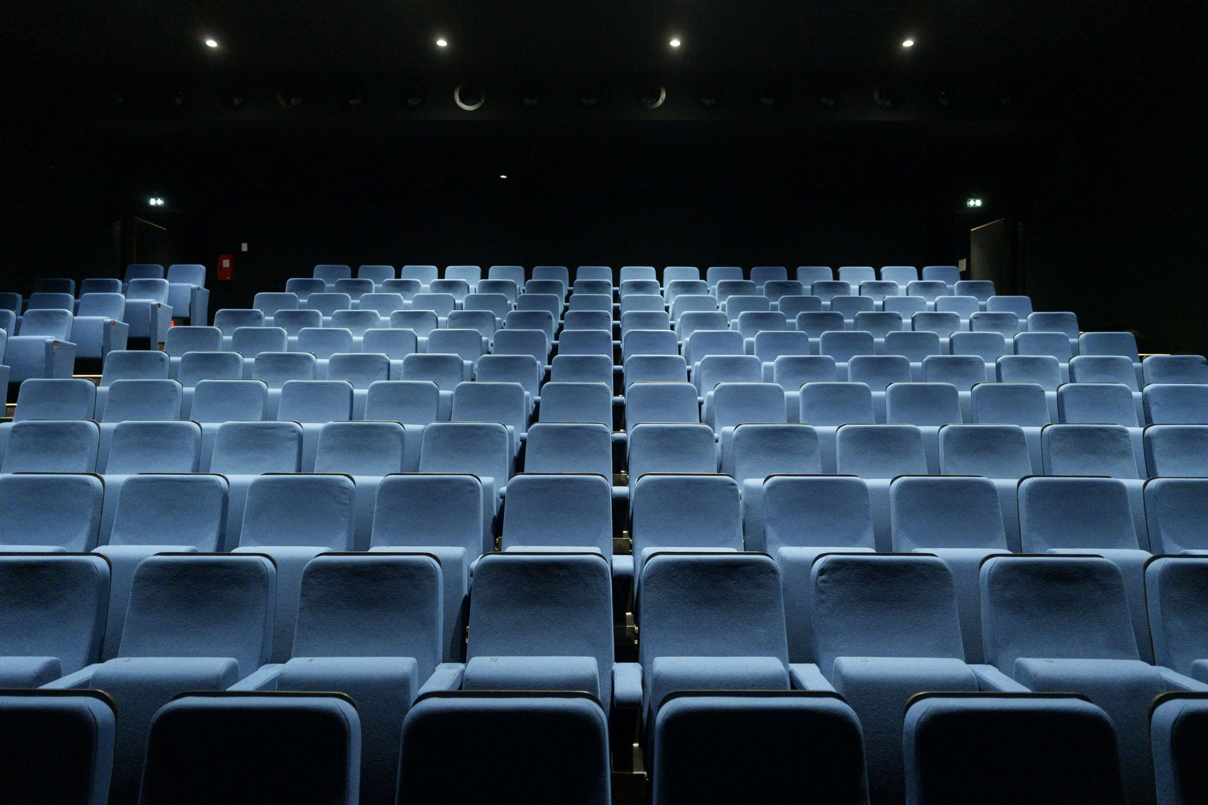 A theater with rows of empty blue upholstered seats facing a darkened stage.