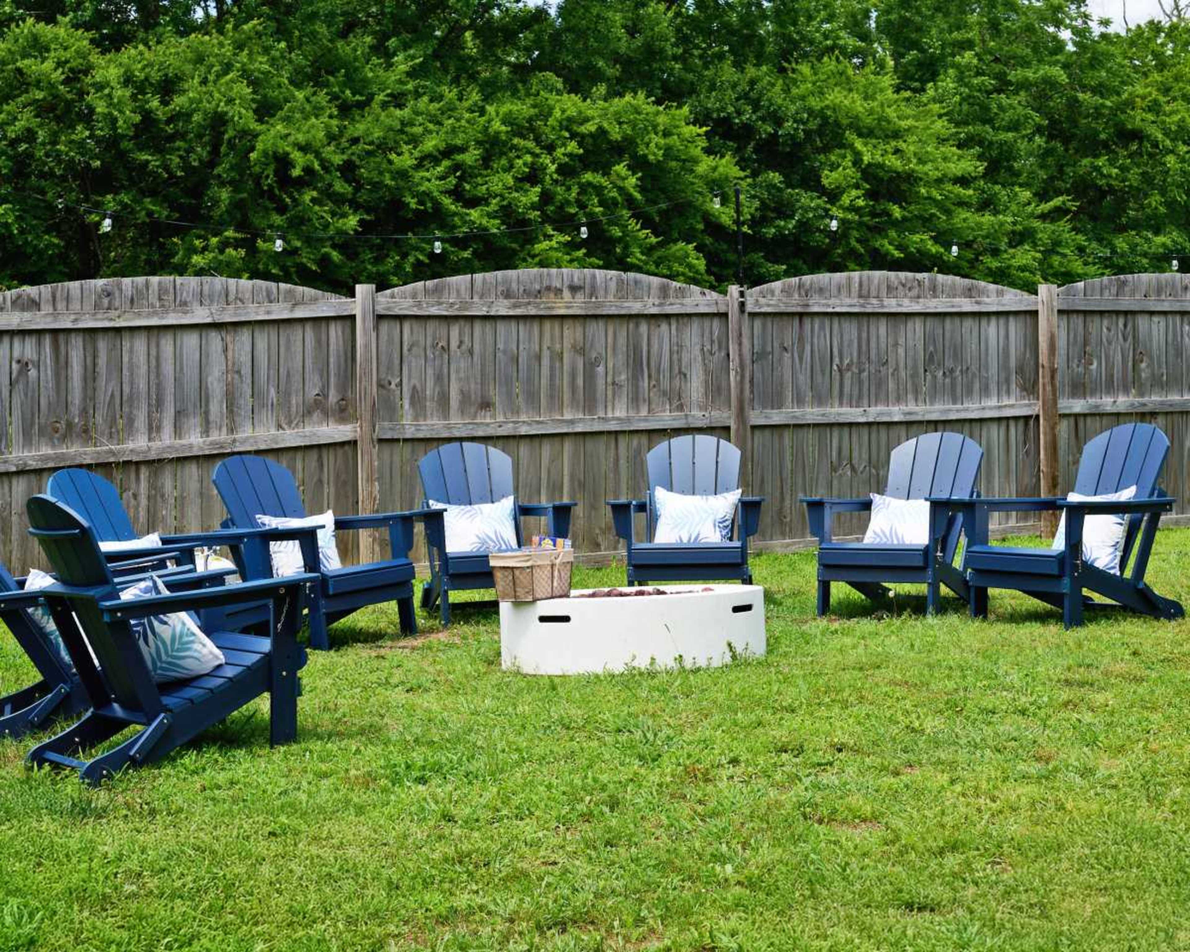 The image shows a grassy area surrounded by a wooden fence featuring six blue Adirondack chairs arranged around a rectangular white table.