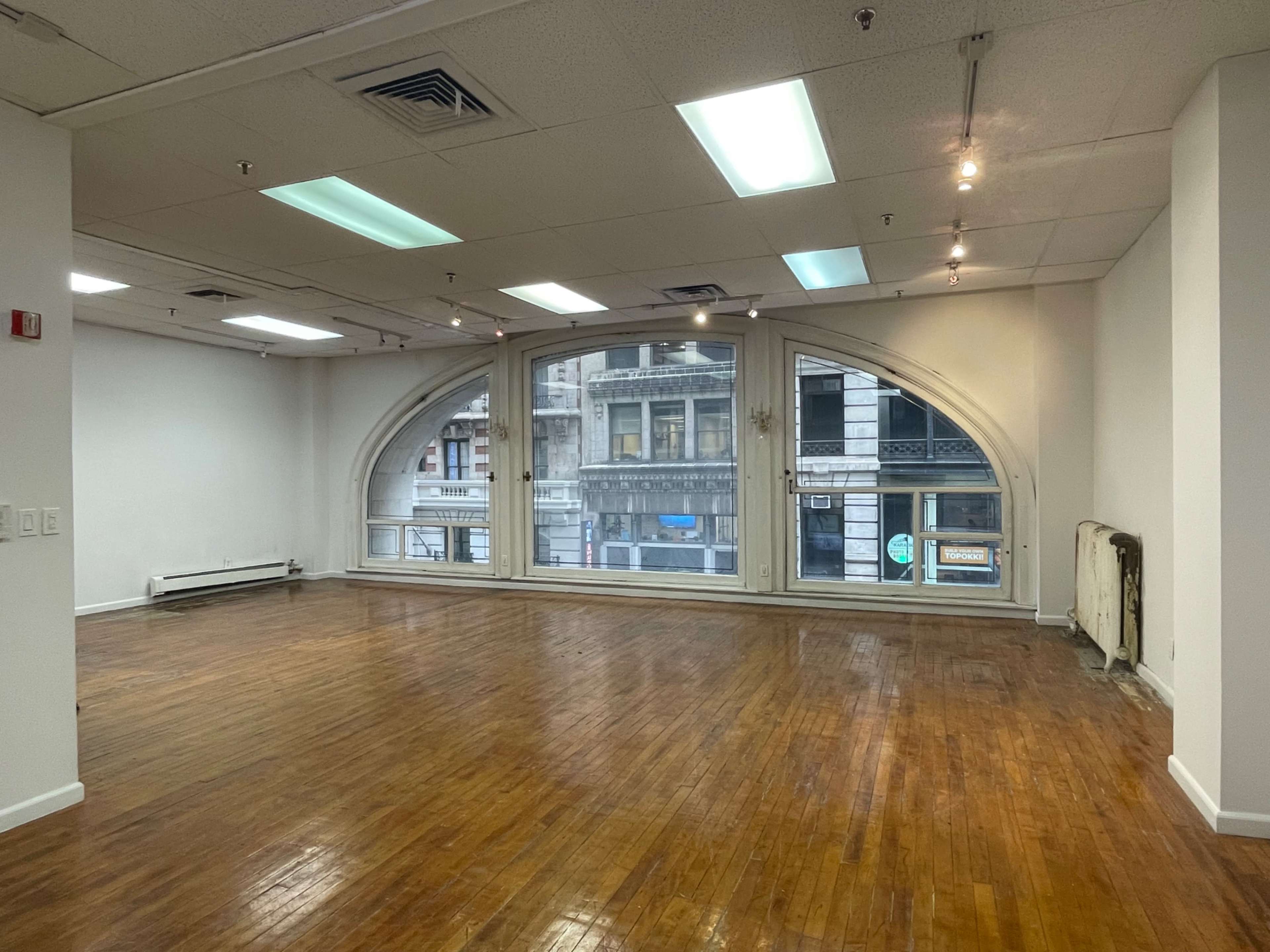 The image shows an empty room with wooden floors and large arched windows overlooking a city street.