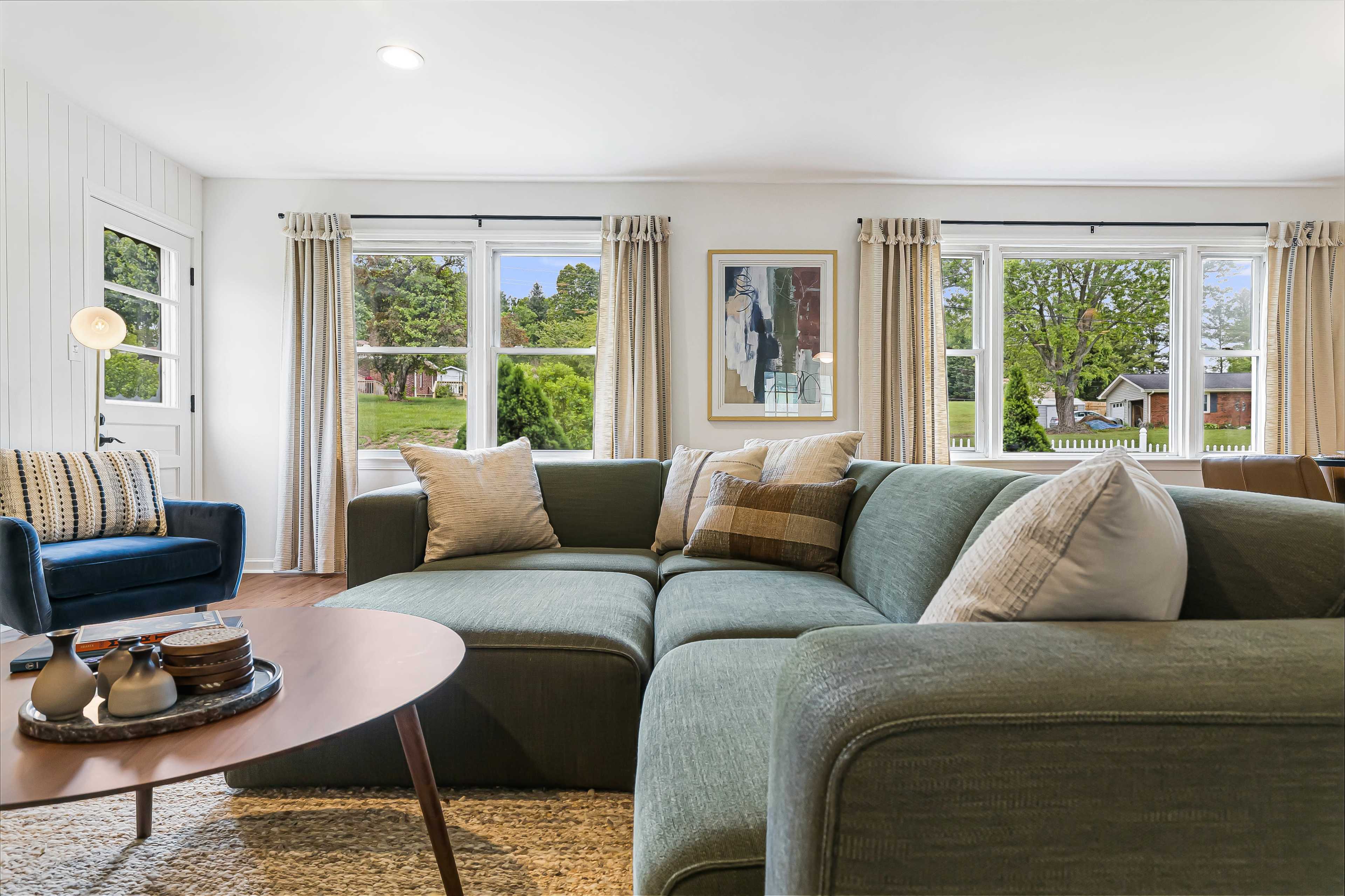 A modern living room features a sectional sofa, a round coffee table with decorative items, and large windows that let in natural light.