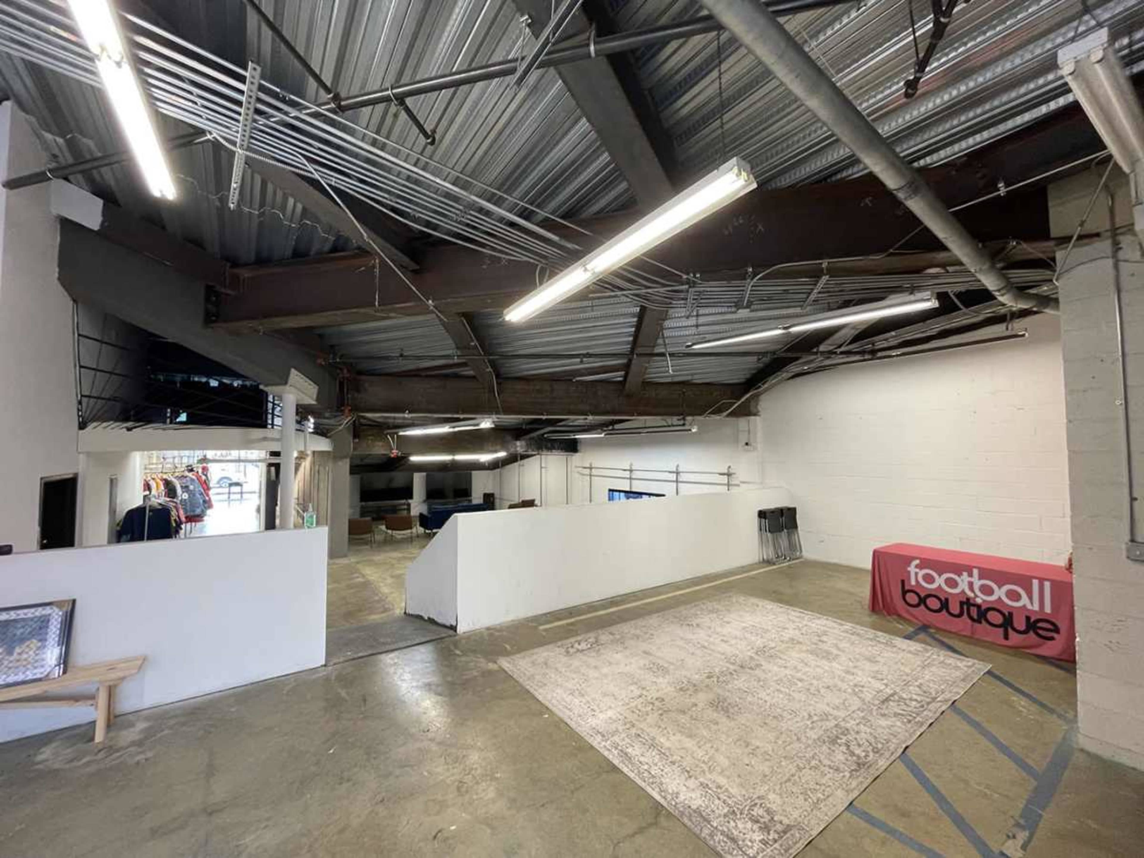 The image shows a spacious interior of a retail space with exposed metal beams, overhead lighting, and a section featuring a promotional table for a football boutique.