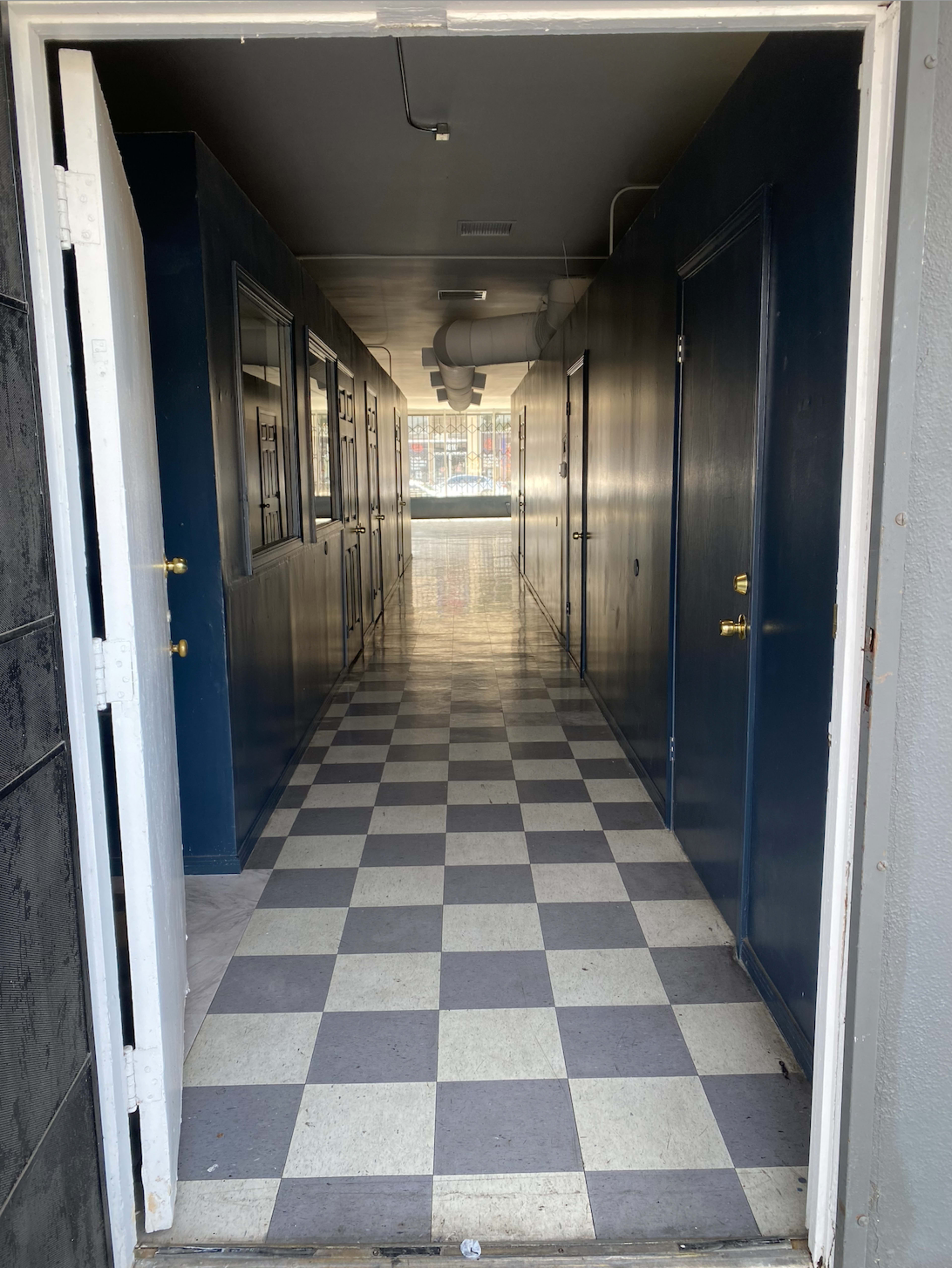 The image shows a hallway with a checkered tile floor and blue walls, leading to a glass door at the end.