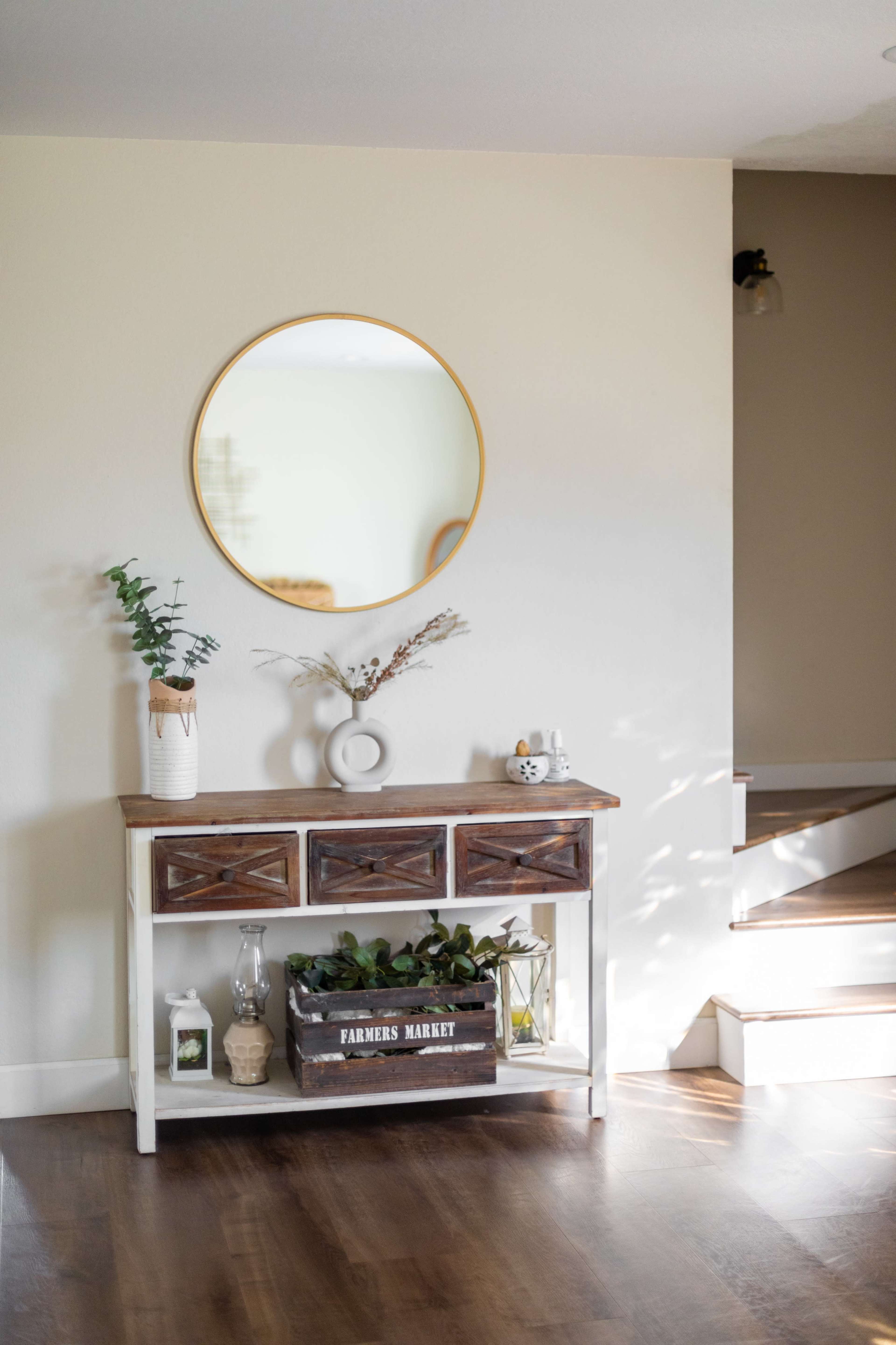 A wooden console table with three drawers and a decorative mirror hangs above it, displaying a vase and a wooden crate labeled "FARMERS MARKET" on a hardwood floor.