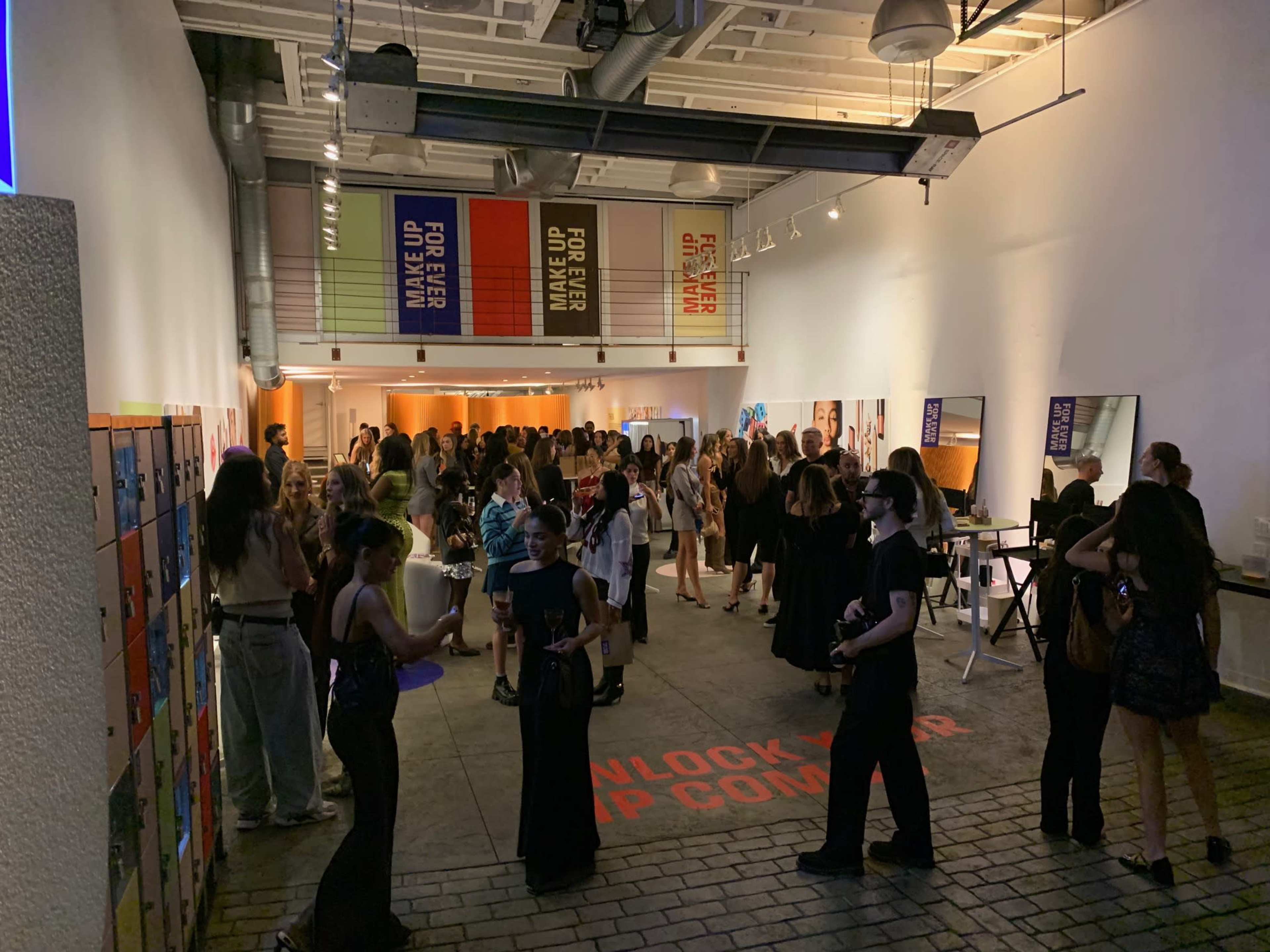 A large group of people mingles in an art gallery, with colorful banners hanging overhead and mirrors set up along one side.