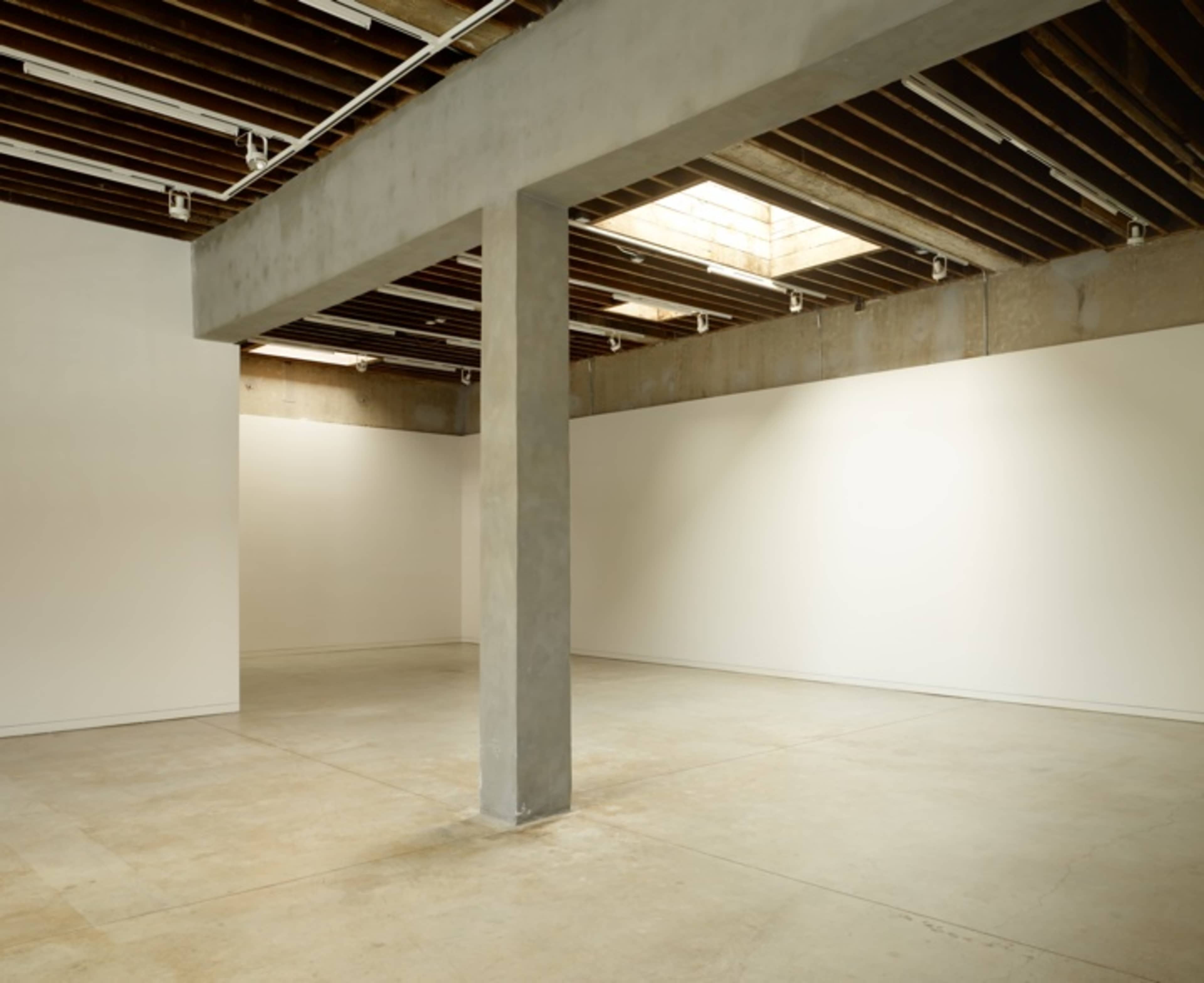 An empty gallery space features plain white walls, a concrete floor, and a central support column.