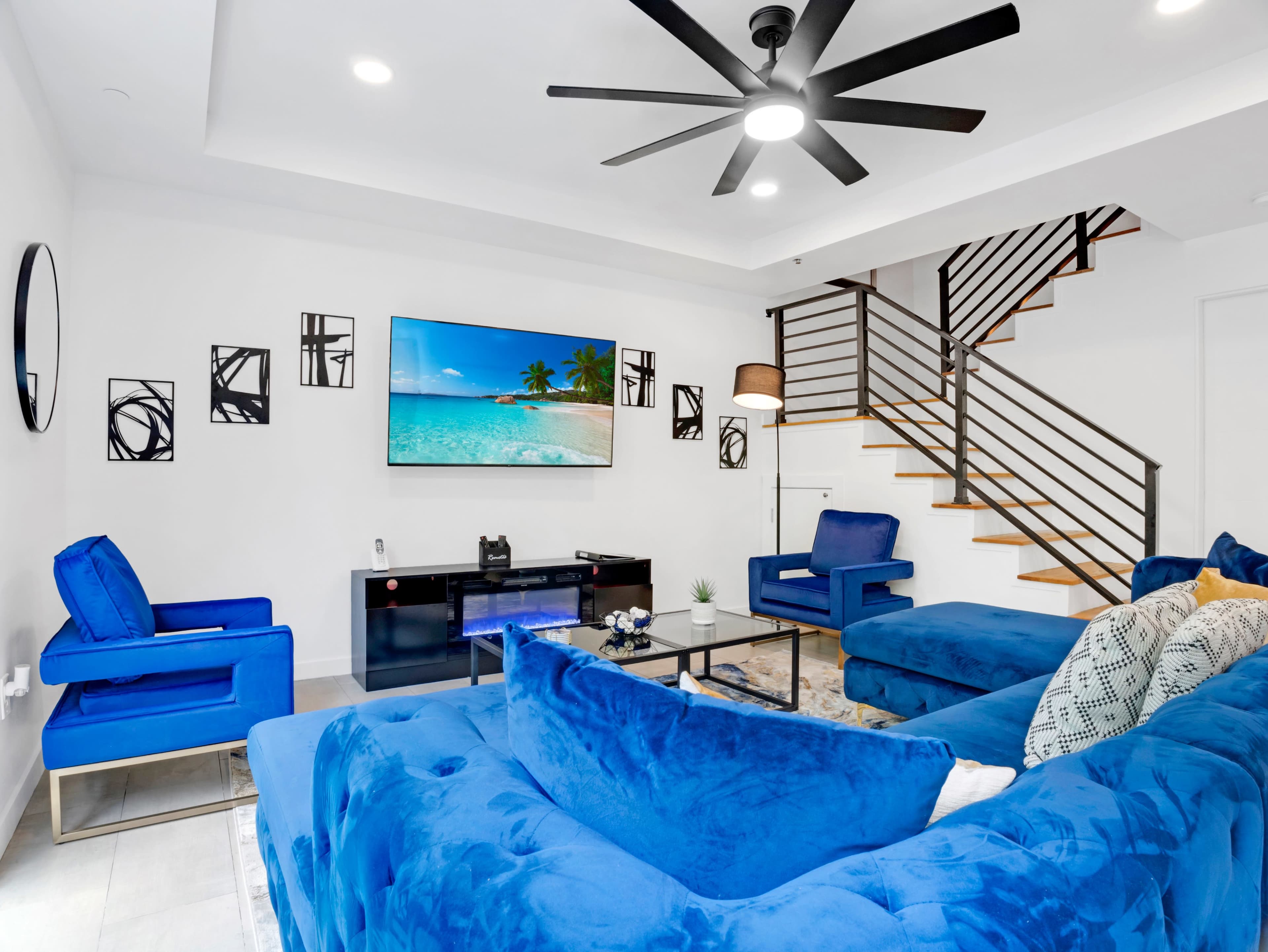 The living room features blue upholstered furniture, a large TV displaying a beach scene, and a staircase leading to an upper level.