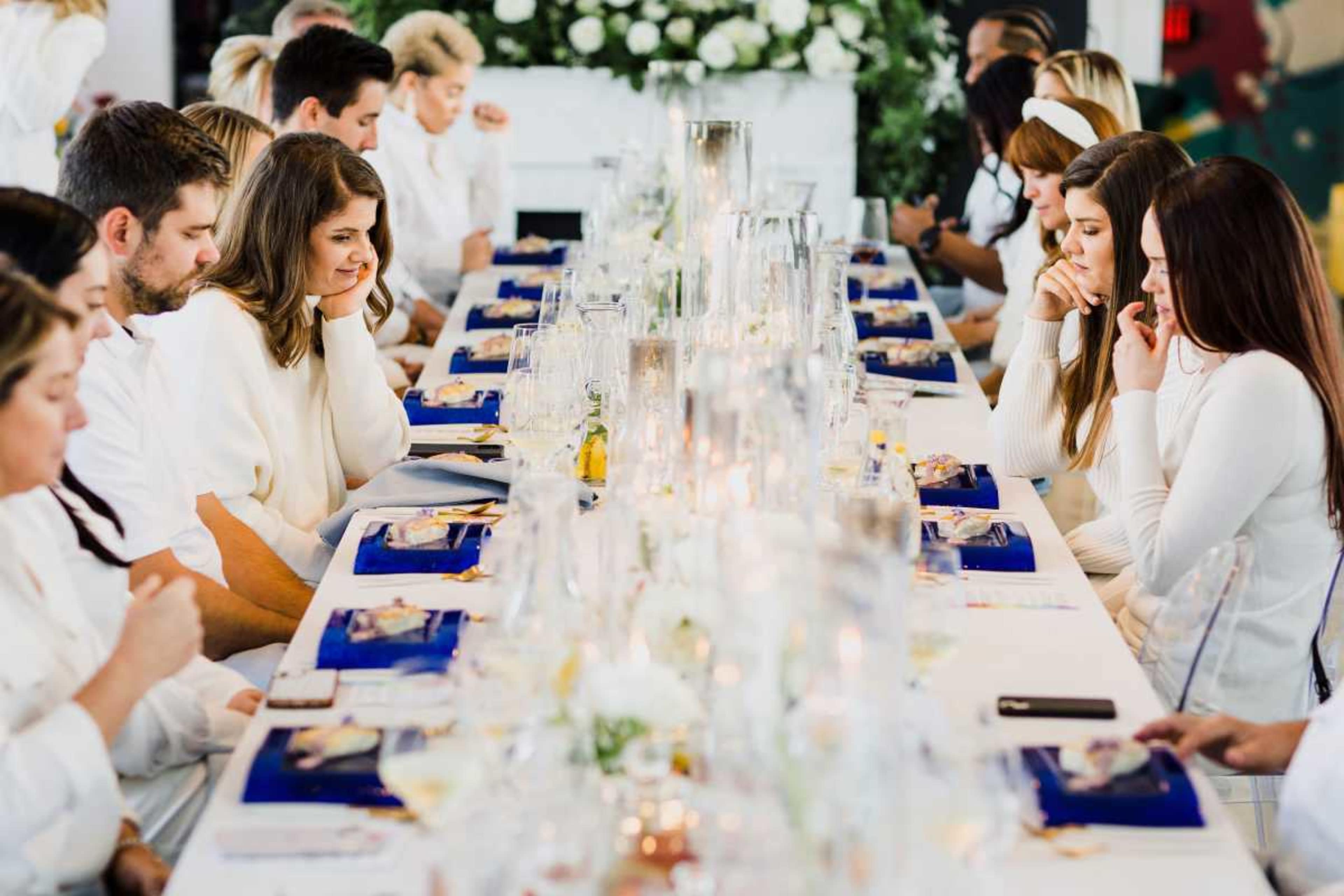 A group of individuals dressed in white sits at a long table set with glasses and menus, engaged in conversation during a dining event.