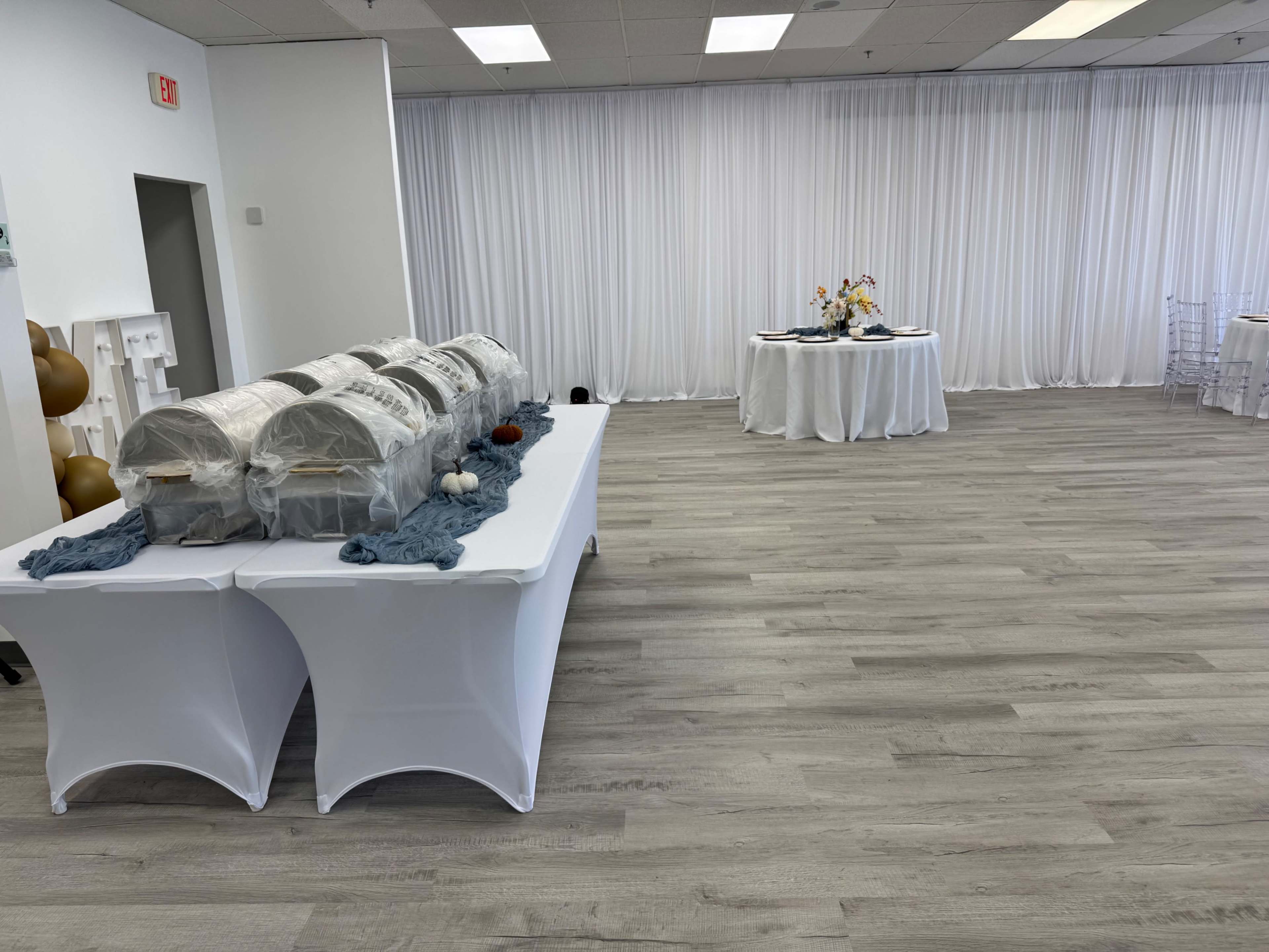 The image shows a spacious interior setup for an event, featuring tables covered with white tablecloths, a display of food under covers, and a decorated table in the background.