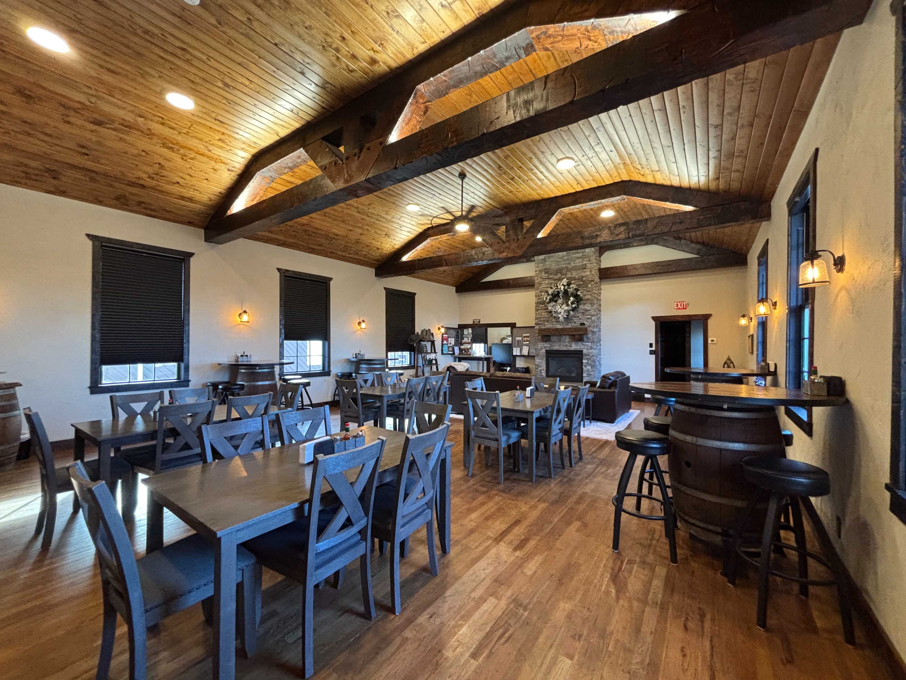 The image shows a spacious dining area with wooden tables and chairs, a stone fireplace, and a bar area, all under a vaulted wooden ceiling.