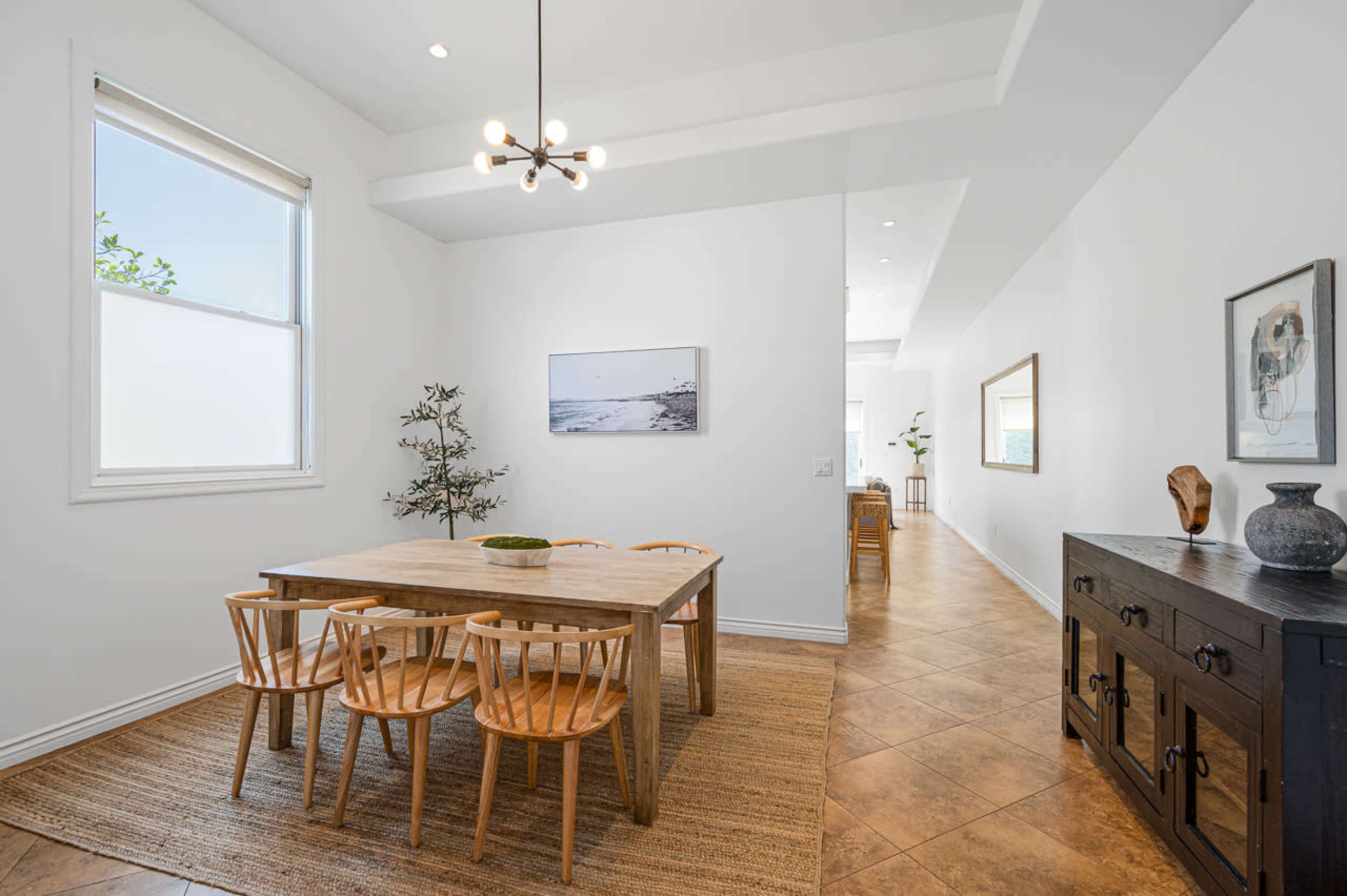 The image shows a dining area with a wooden table and chairs, a plant, and a framed picture on the wall, all set against a light-colored background.