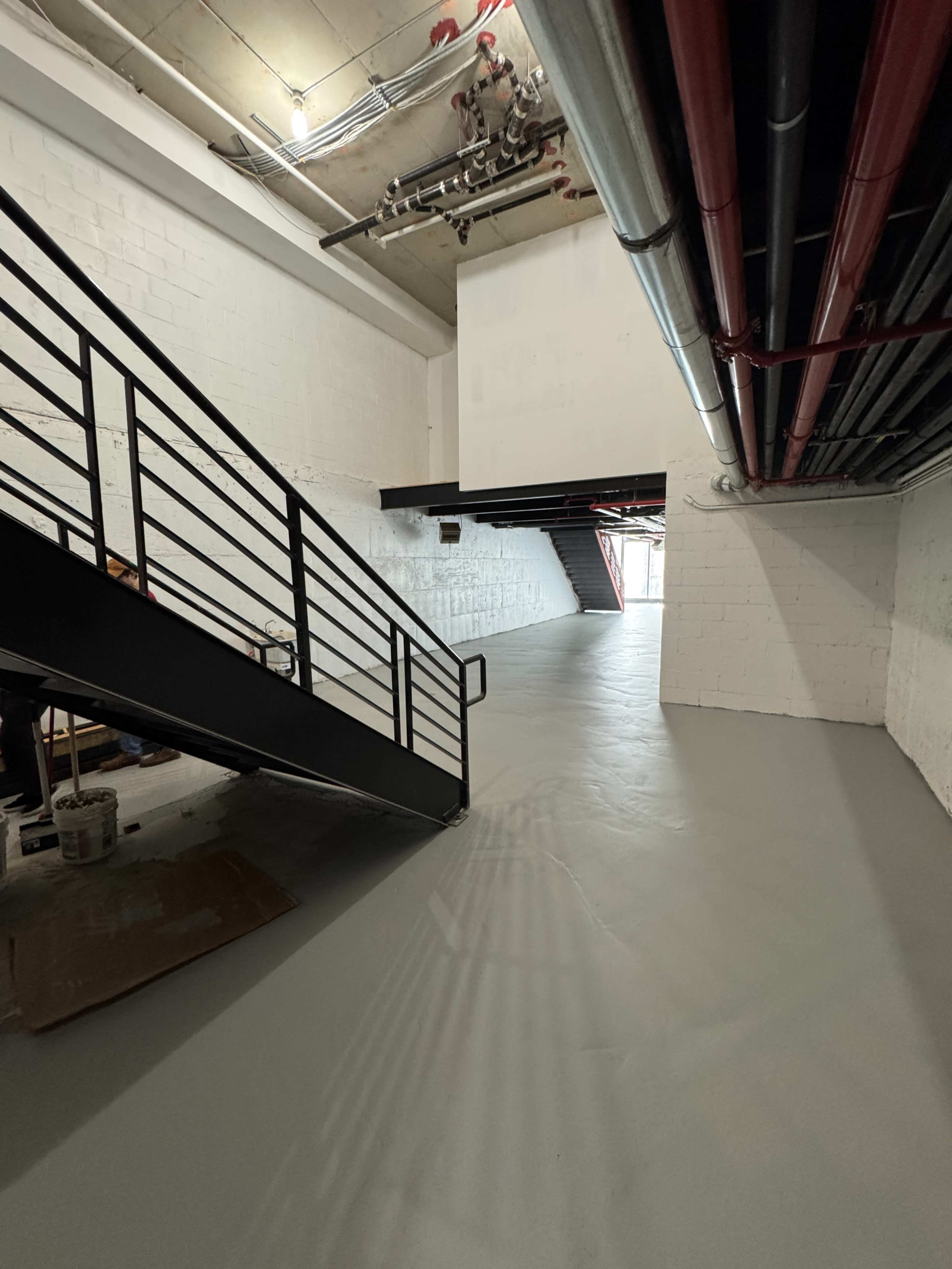 The image shows a gray-floored interior space with a staircase on the left, exposed pipes and ductwork above, and an open archway in the background.