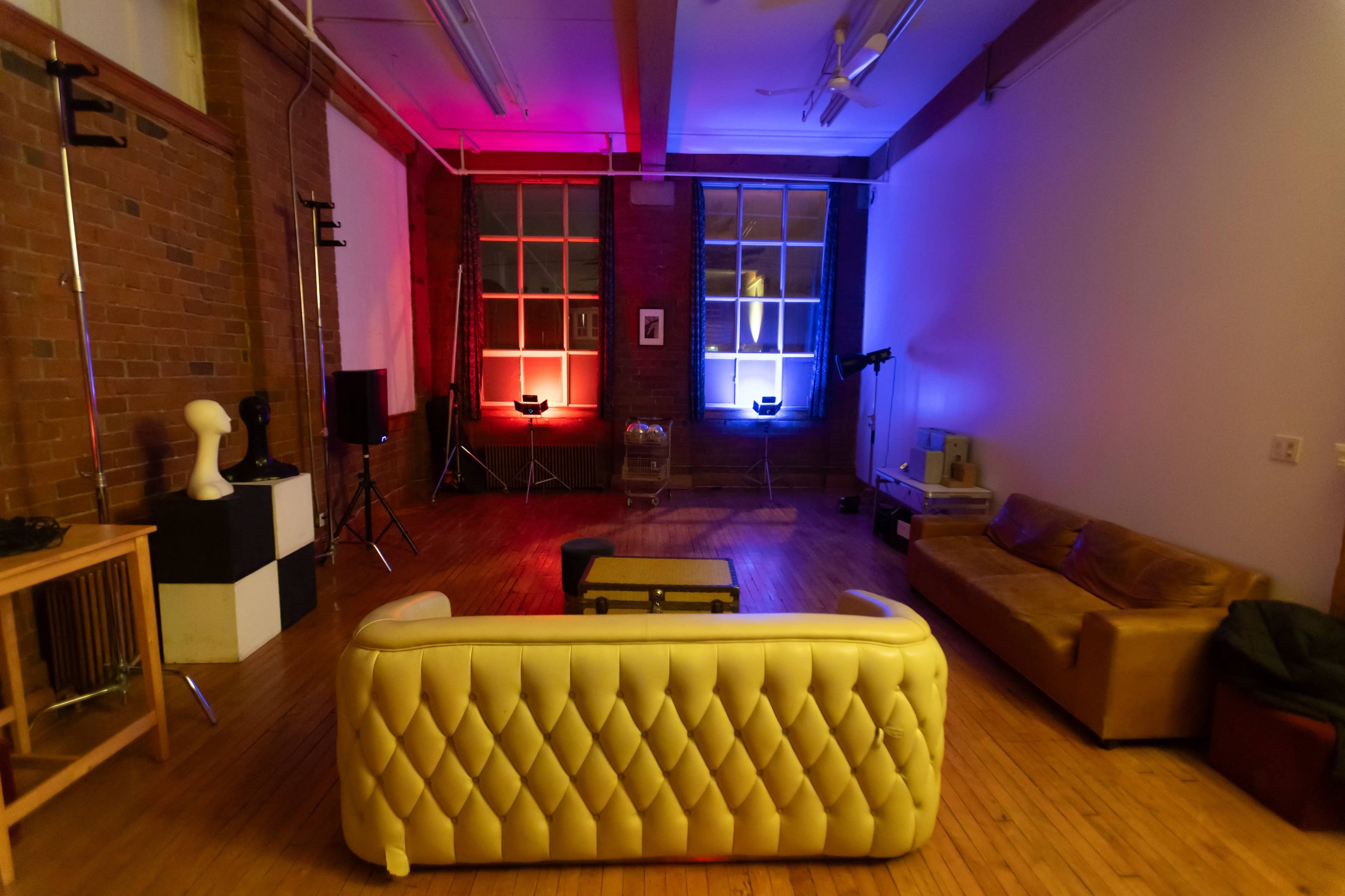 The image shows a spacious room with brick walls, featuring a yellow leather couch, a coffee table, and various lighting equipment arranged around two windows illuminated in red and blue.